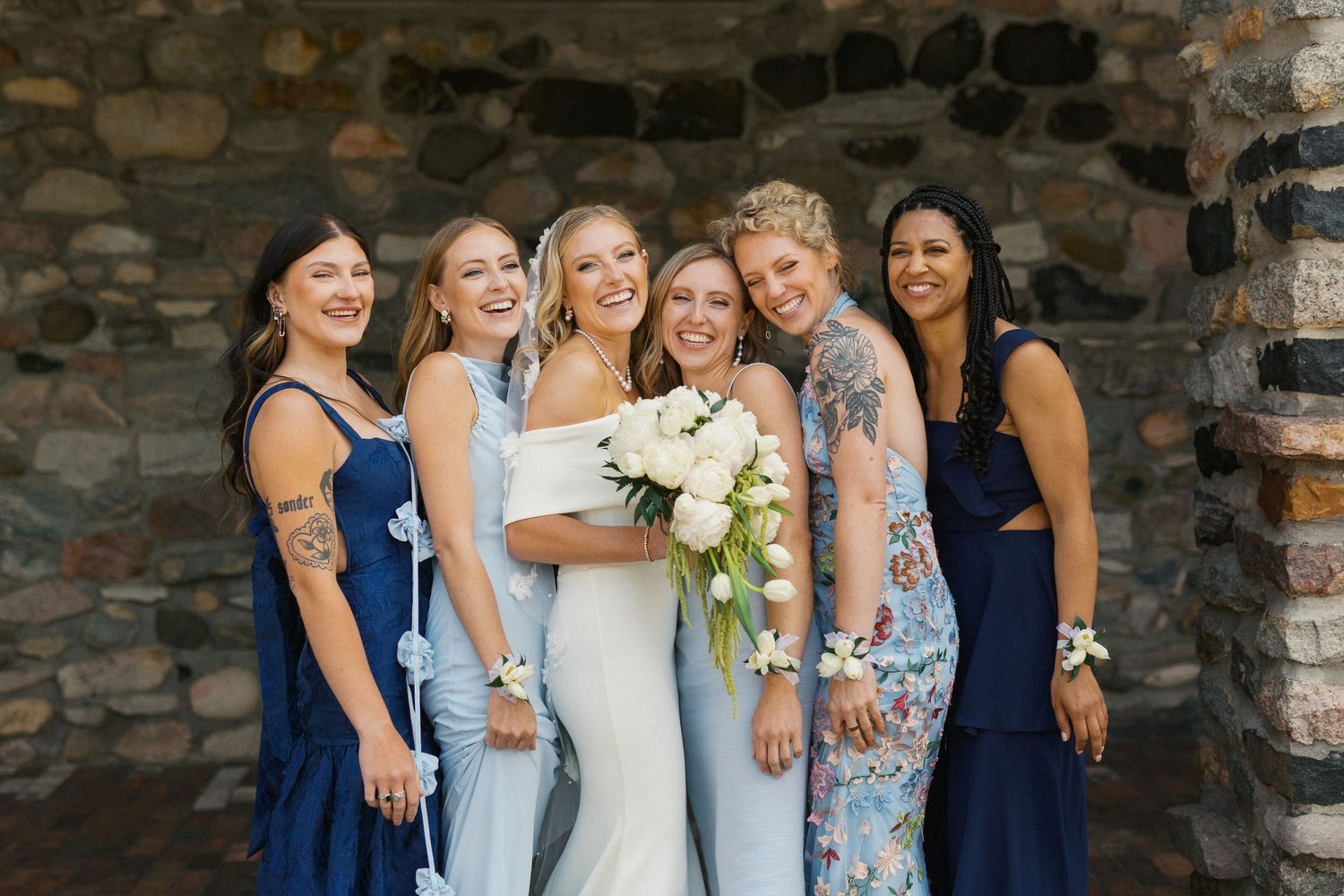 A bridal party in mixed blue dresses poses together against a stone wall backdrop.