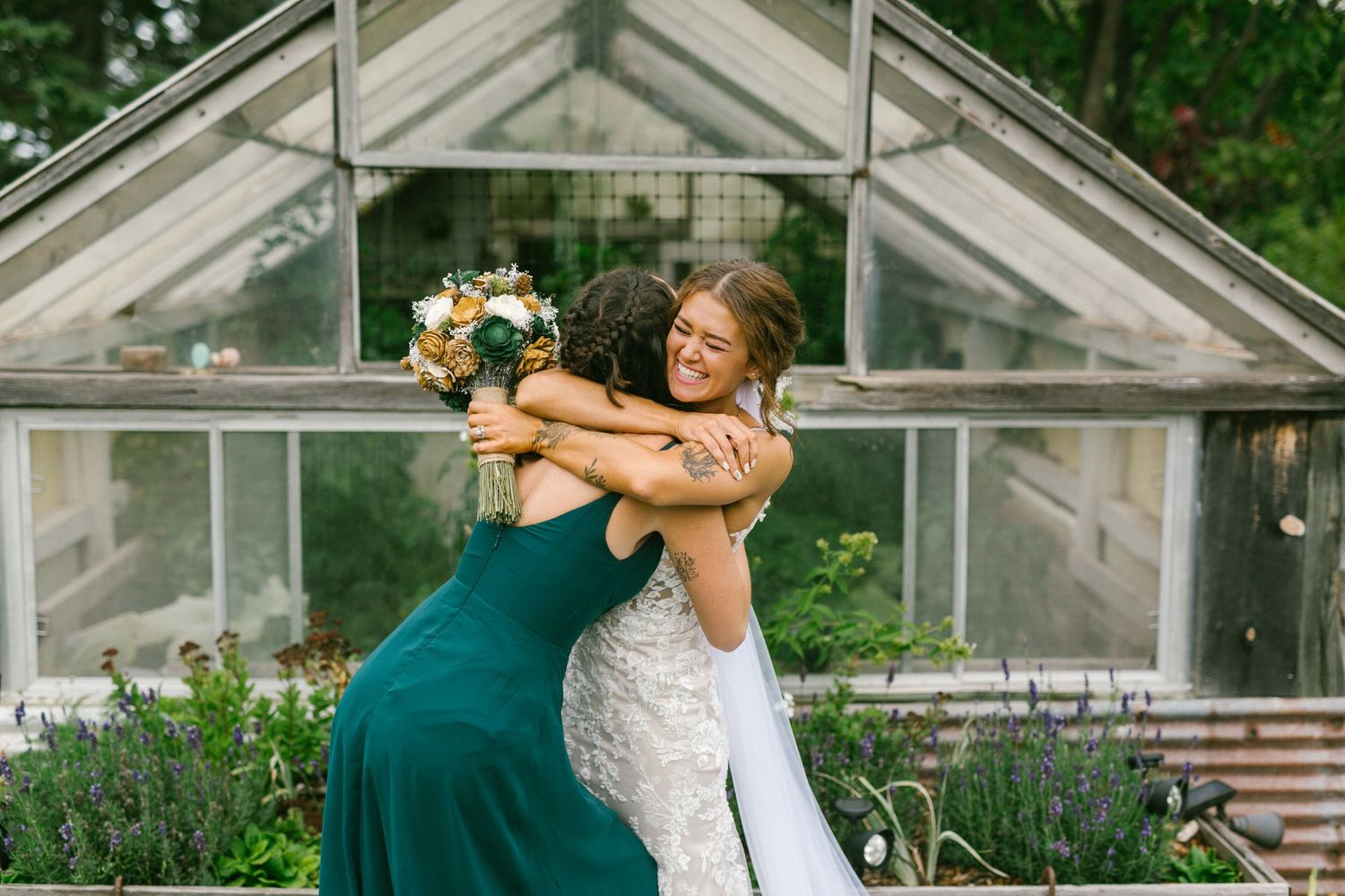 Two people in formal dresses share a joyful embrace and laugh in front of a glass greenhouse with flowers.
