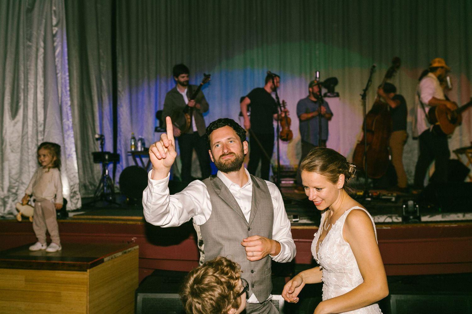 Wedding guests dance together on a wooden dance floor with a live band performing on stage in the background.