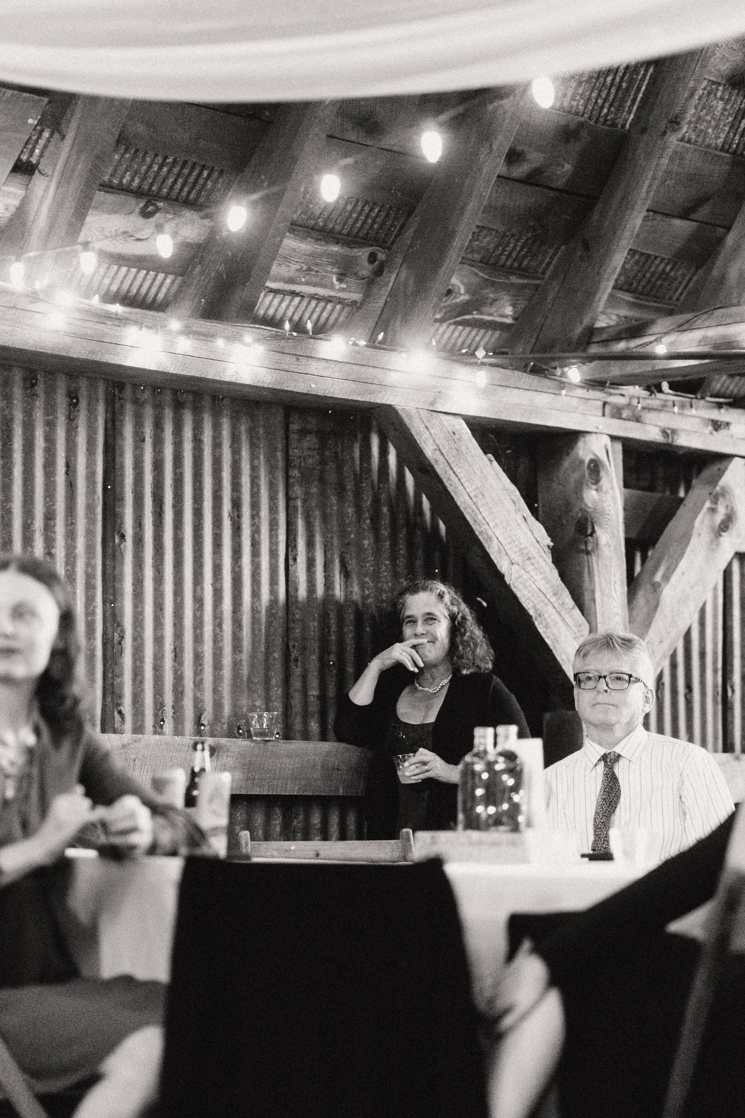 Black and white photo of guests enjoying a celebration in a rustic barn setting with string lights.
