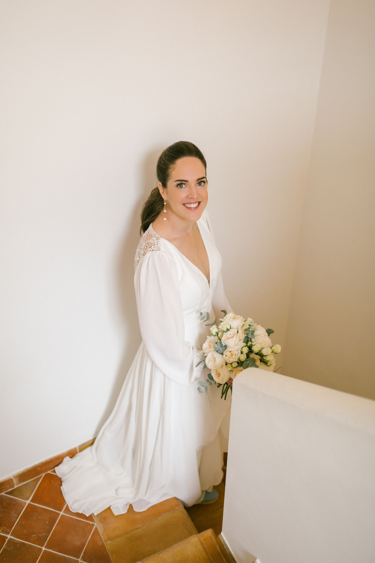 A bride wearing an elegant long-sleeved white wedding dress with lace details holds a soft pastel bouquet on stairs.