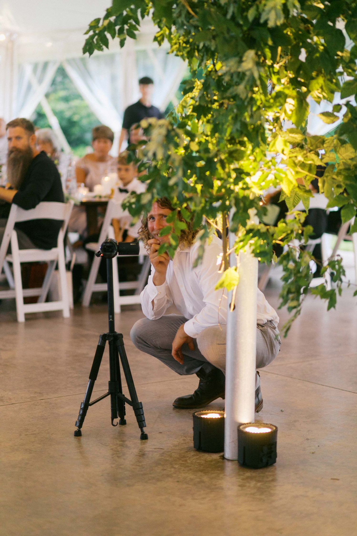 White candles illuminate a rustic outdoor reception setting under tree branches at dusk.