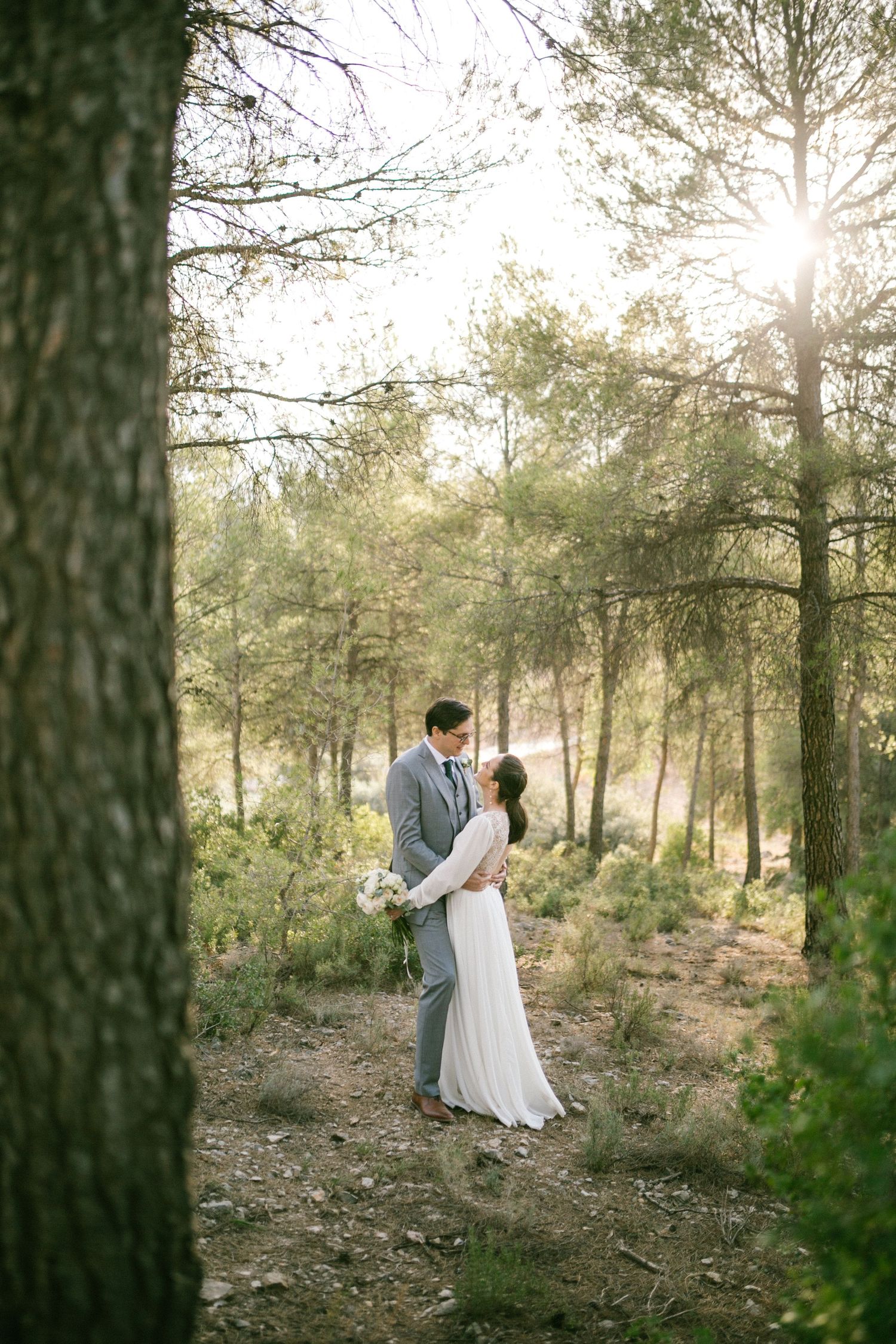 A couple embraces in a sun-dappled forest setting with pine trees and natural light filtering through the branches.