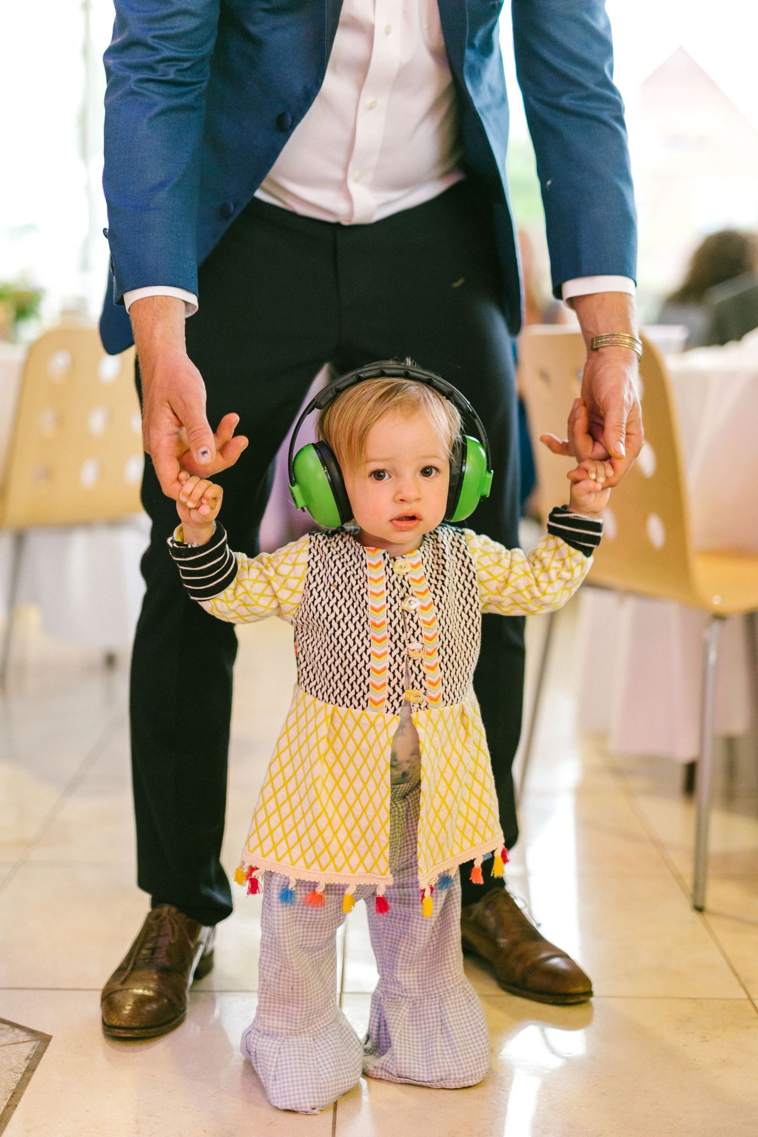 Small child wearing yellow dress and headphones standing between adult legs at formal event.