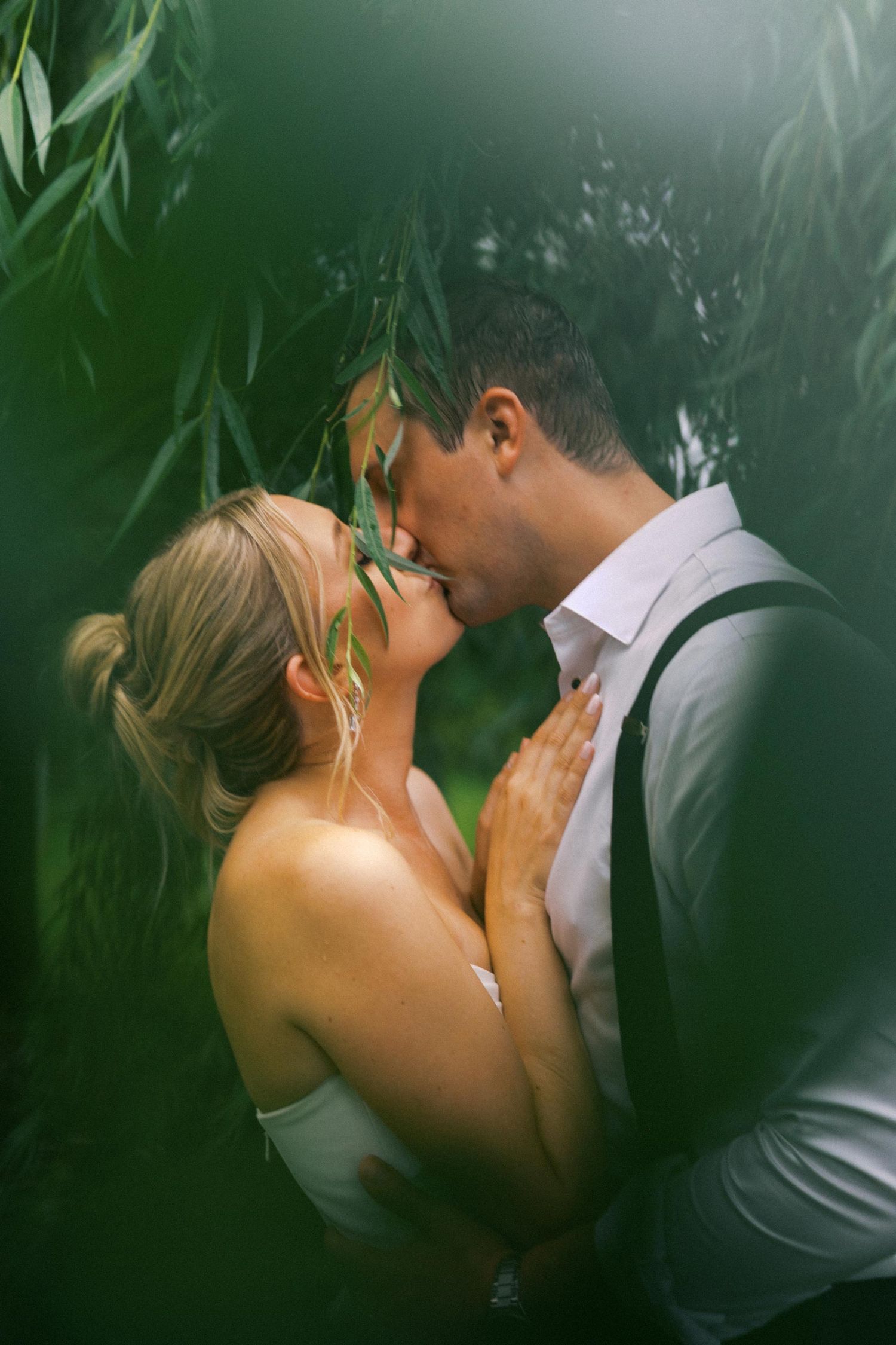 A romantic couple shares an intimate kiss in front of a lush green garden and inside a willow trees branches.