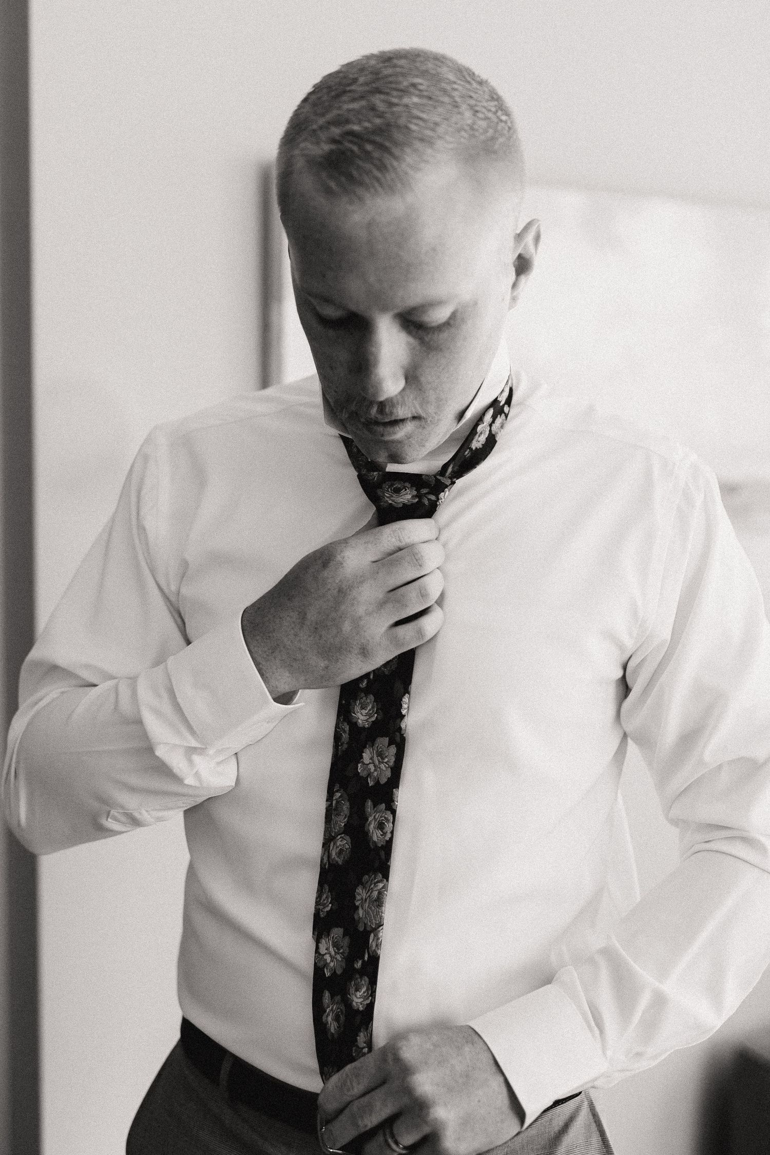 A person in a white shirt adjusts their dark patterned tie in a black and white photo.