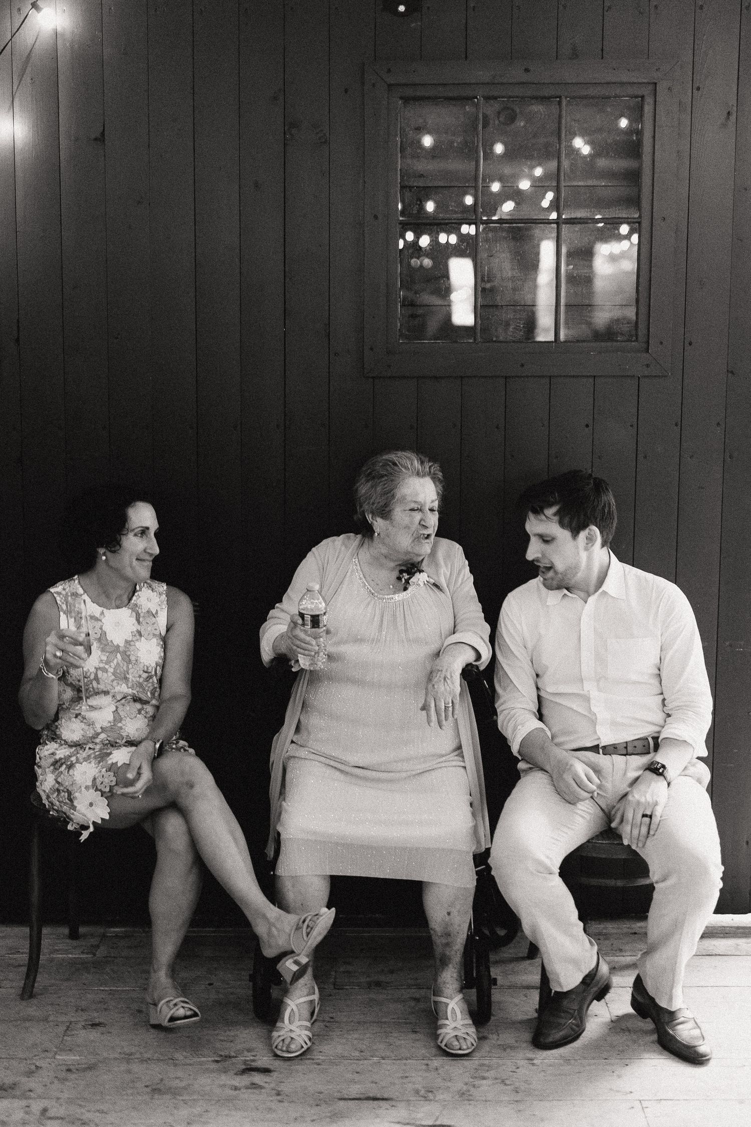 Black and white photo of two women and a man sitting outdoors at a wedding reception near a rustic barn.