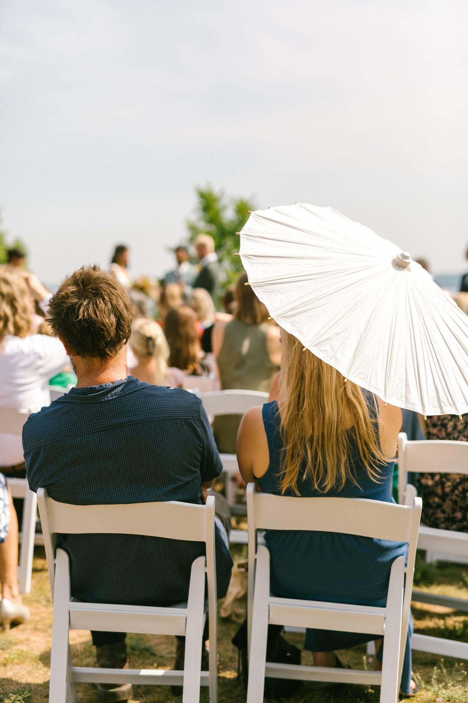Wedding guests seated in white chairs with paper parasols at an outdoor ceremony.