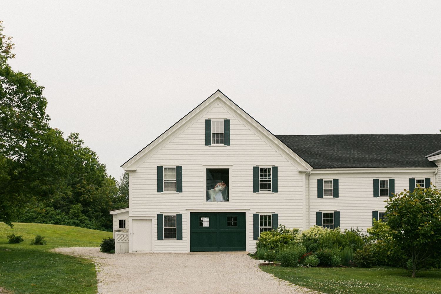 White colonial farmhouse with green shutters and garage doors, surrounded by lush greenery on an overcast day.