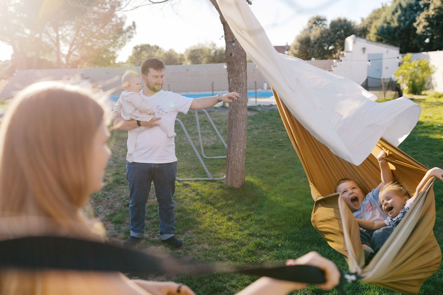 Family enjoying a sunny backyard with children laughing in a hammock while dad holds baby.