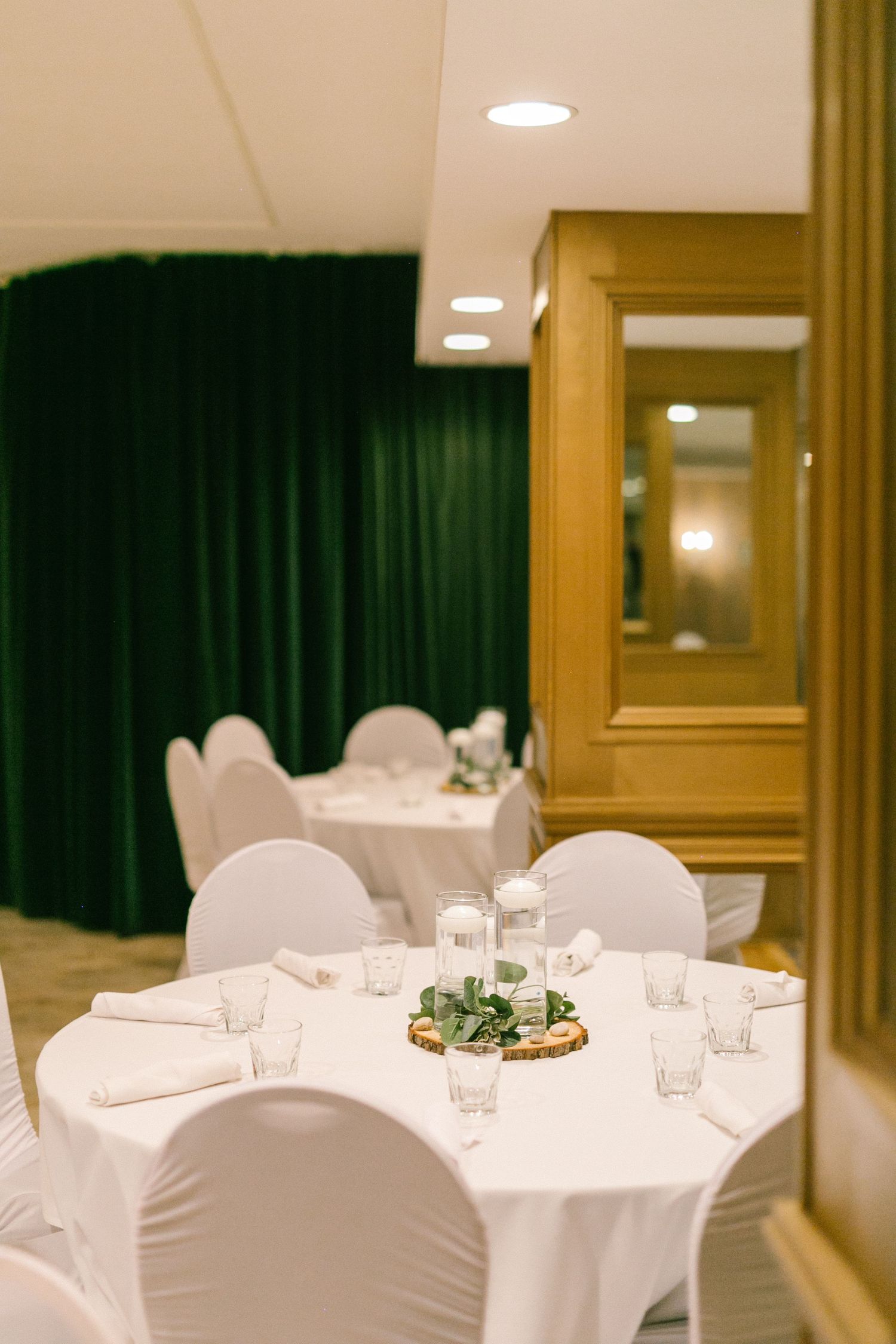 Elegant banquet hall setup with round tables covered in white linens and chairs in a warm-lit room with dark curtains.