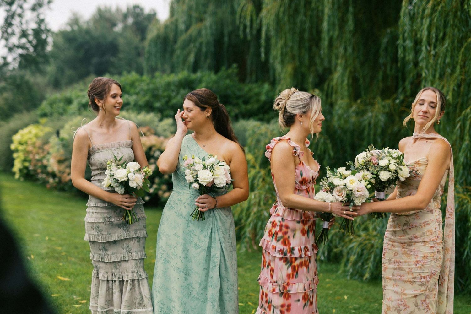 Bridesmaids in floral and pastel dresses hold bouquets of white flowers while feeling emotional a lawn with weeping willows during ceremony.
