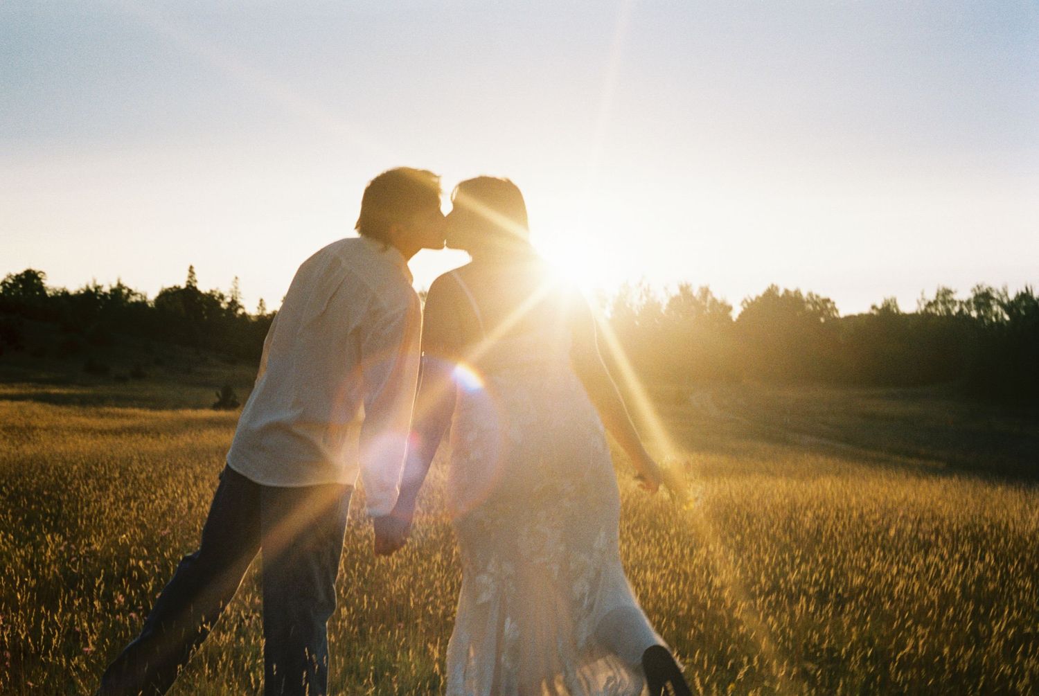 Couple sharing a romantic kiss in a golden sunlit field at sunset with lens flare.