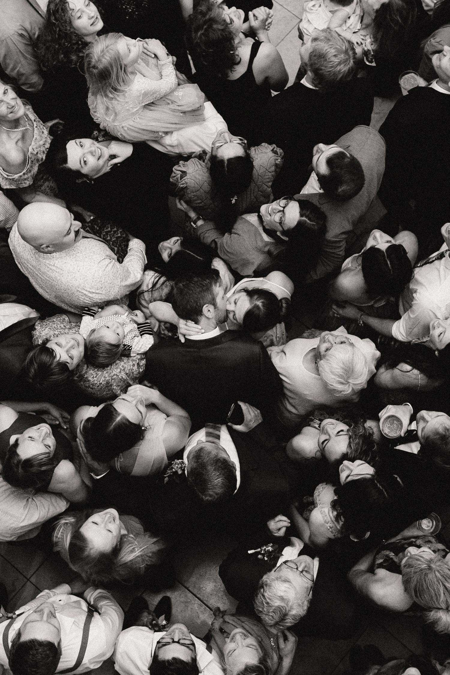 Aerial black and white photo of a crowd surrounding a bride and groom as they kiss.