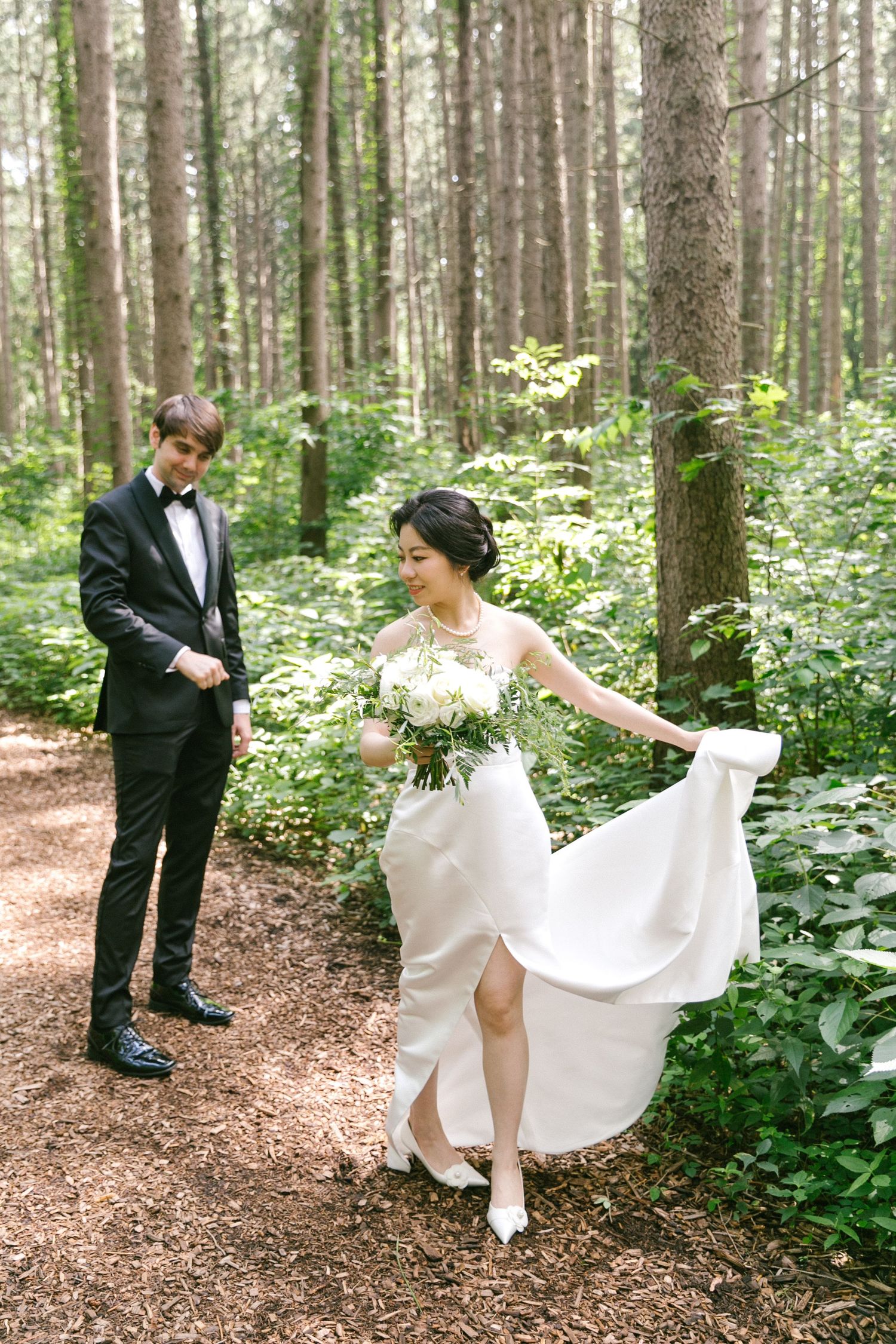 Bride in white gown holds bouquet while groom in tuxedo watches in a lush green forest wedding photo.