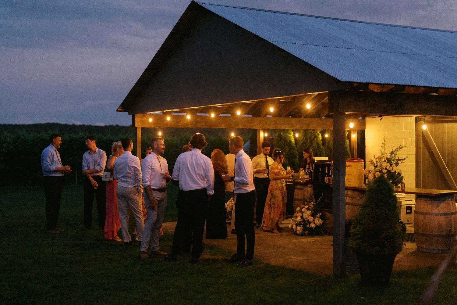 People gather at a rustic barn venue during twilight with string lights creating a warm ambiance at a wedding reception.