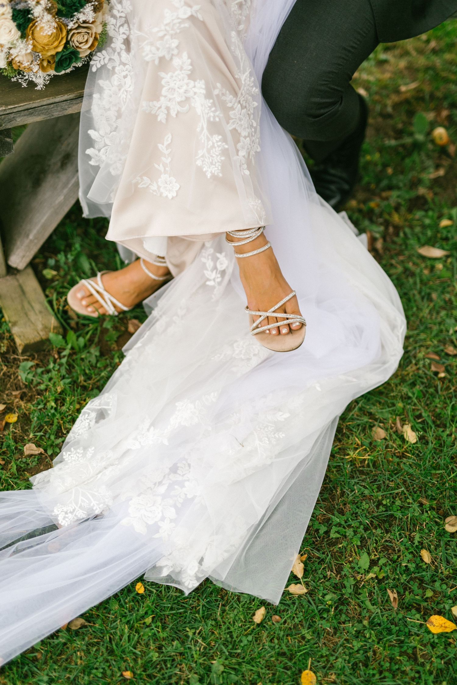 Elegant bridal feet in sparkly sandals peek out from under a flowing white lace wedding dress on green grass.