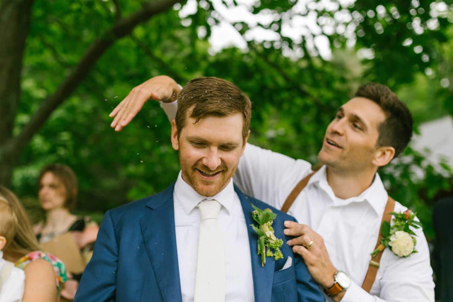 Two people in wedding attire laughing together outdoors with greenery in the background.