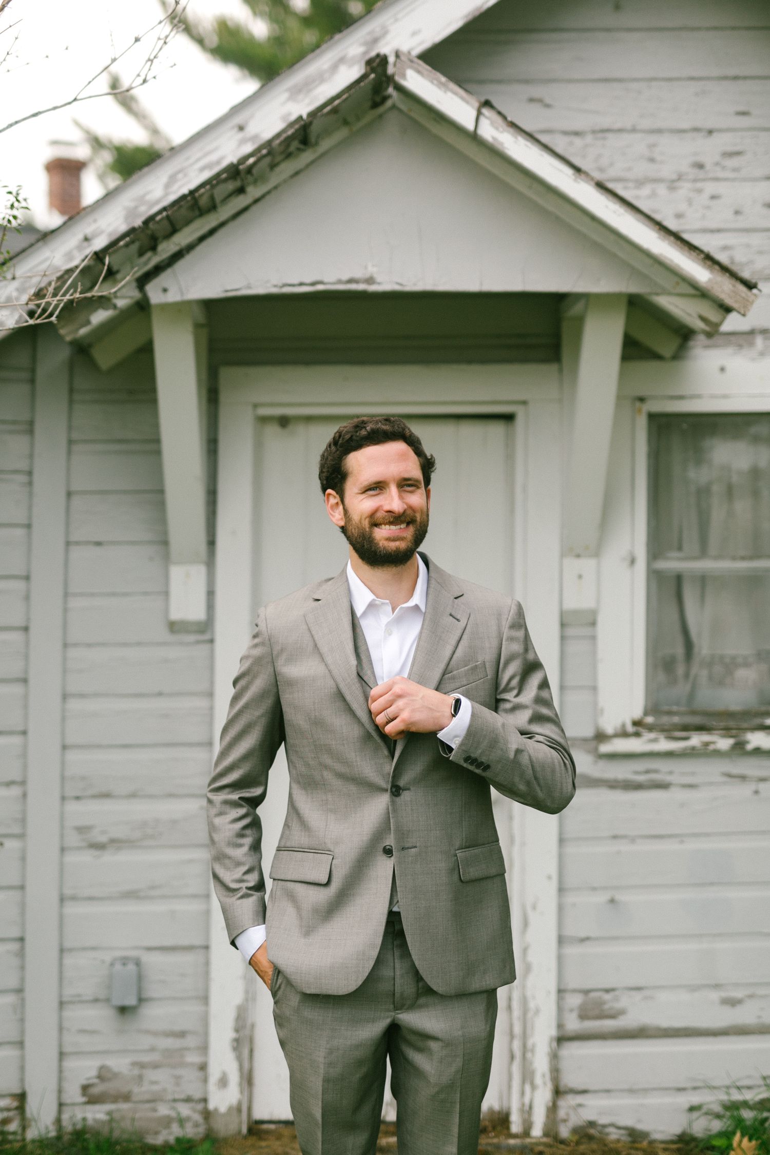 A person wearing a light gray suit and white shirt stands smiling in front of a white farmhouse exterior.