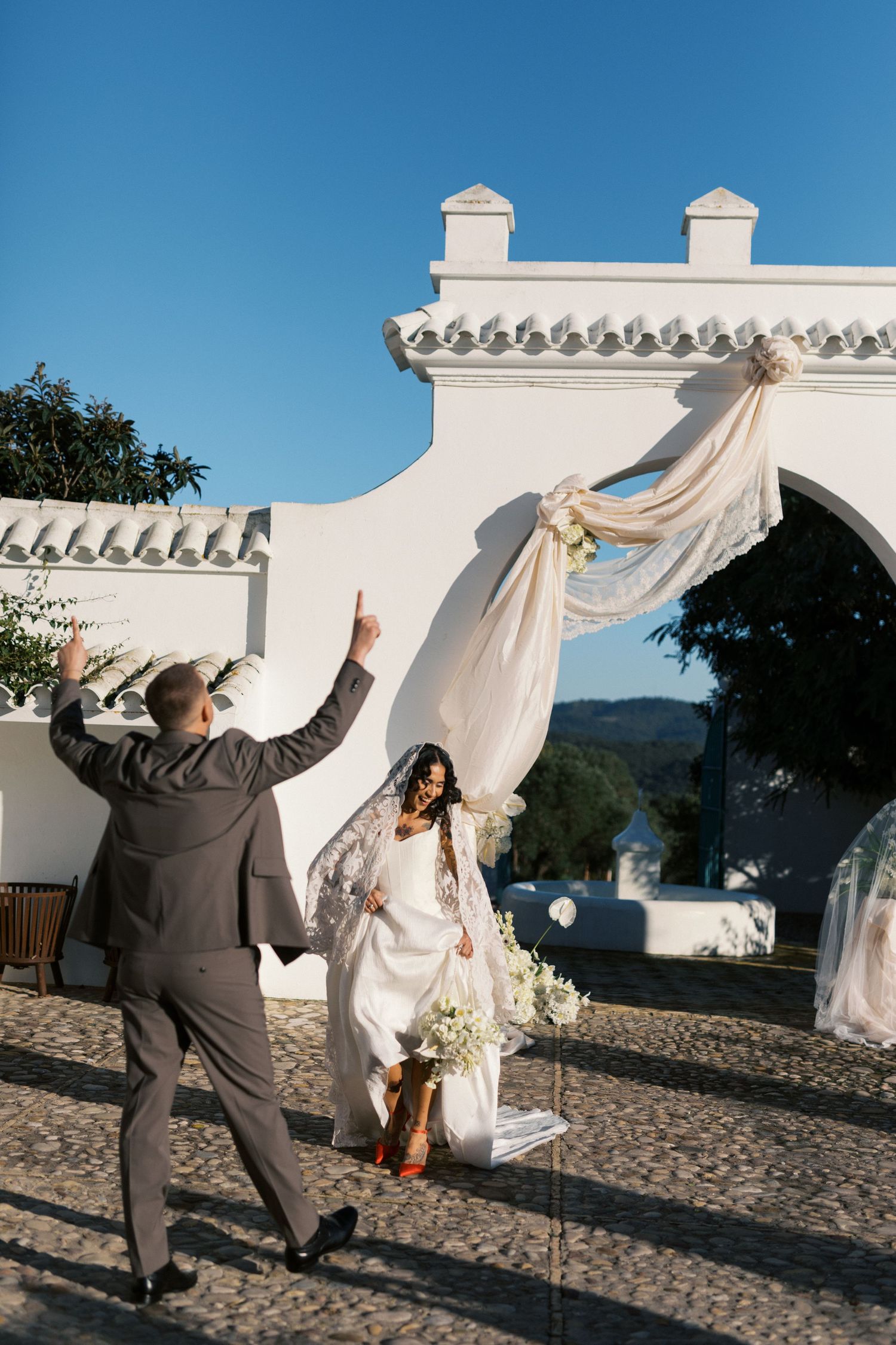 Joyful groom raises hand as bride sits on swing near white arch with flowing fabric at outdoor wedding.