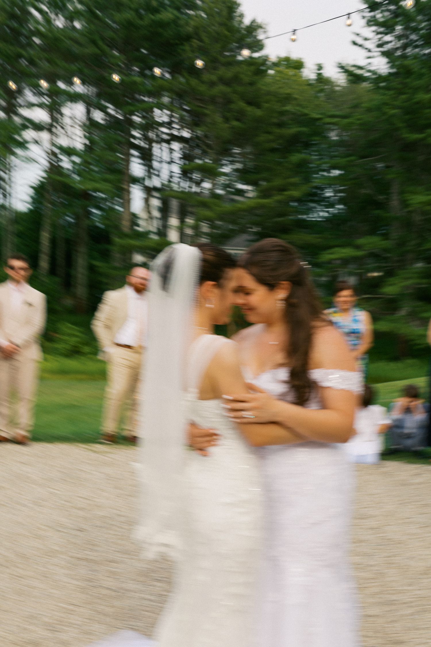 Two brides share a first dance outdoors at a wedding reception surrounded by lush green trees and string lights.