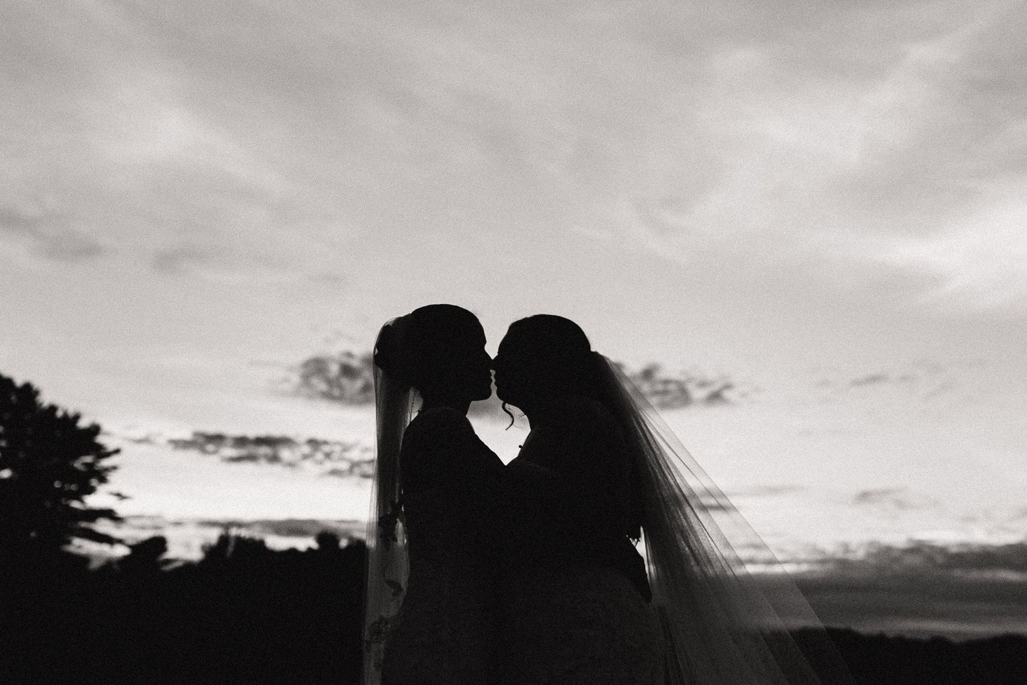 Silhouette of two brides sharing a kiss at sunset, with a flowing veil, in a romantic black and white wedding photo.