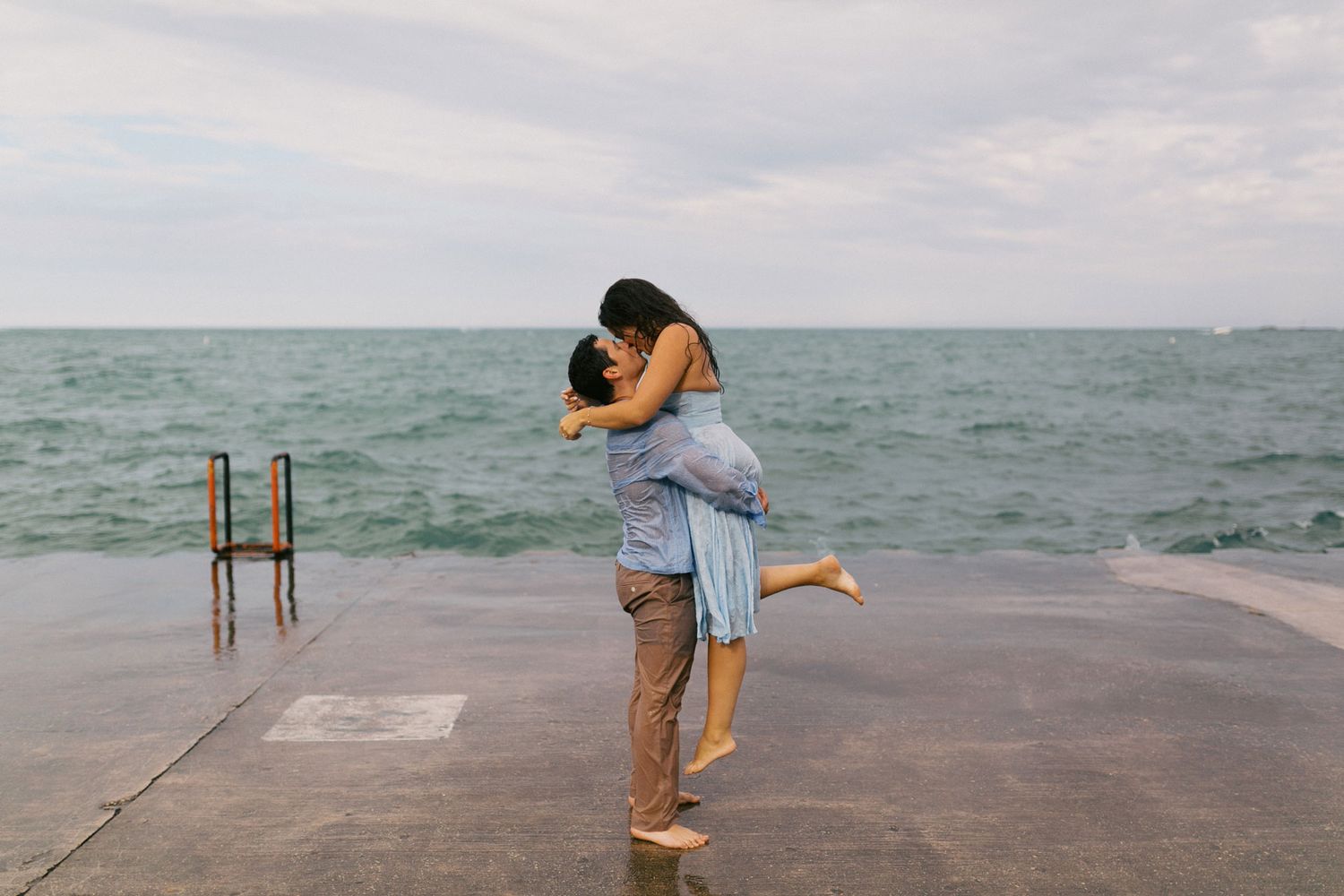 Couple sharing a joyful lift and embrace on a lakeside pier with choppy water behind them.