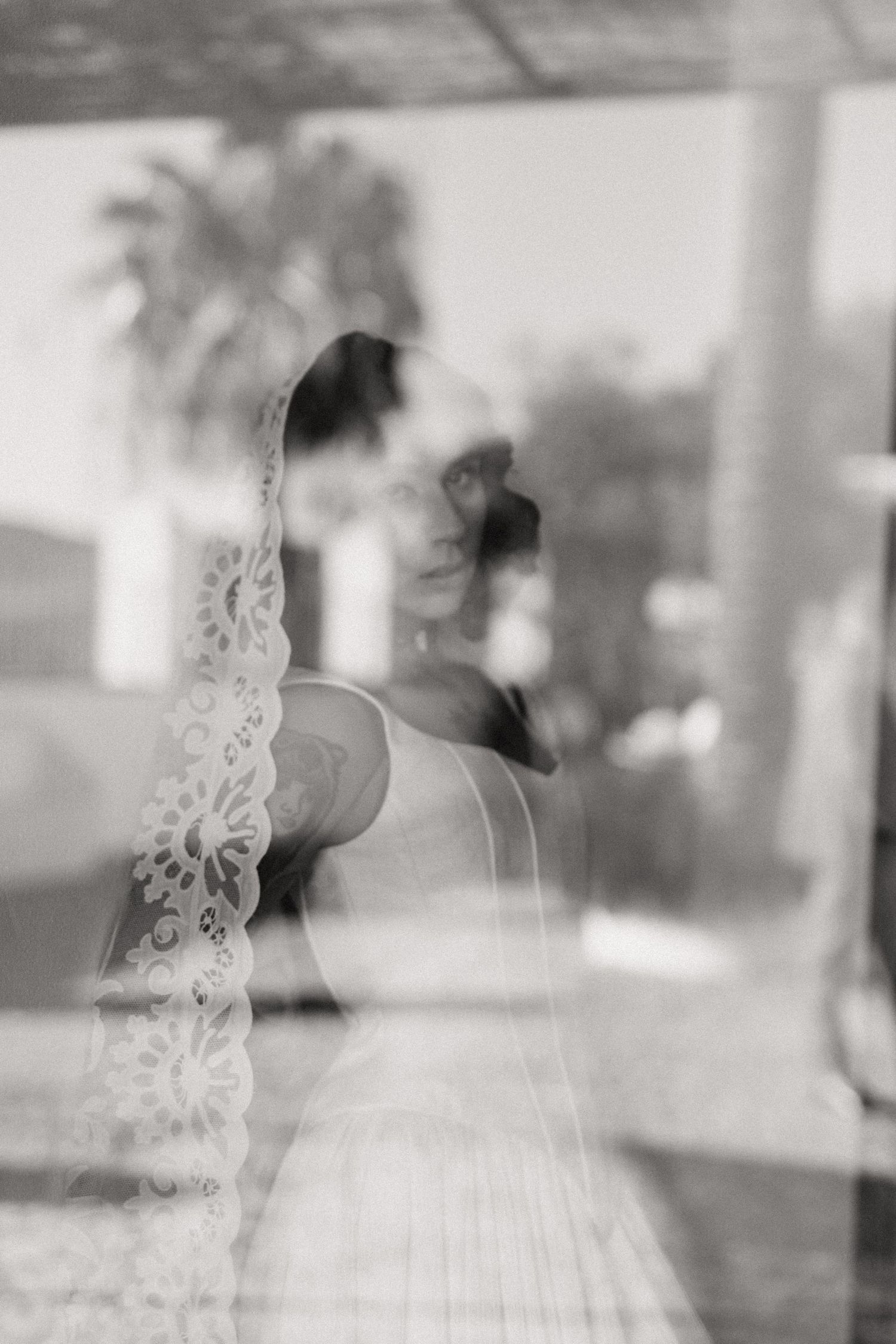 Black and white artistic shot of bride in lace veil reflected through glass window.