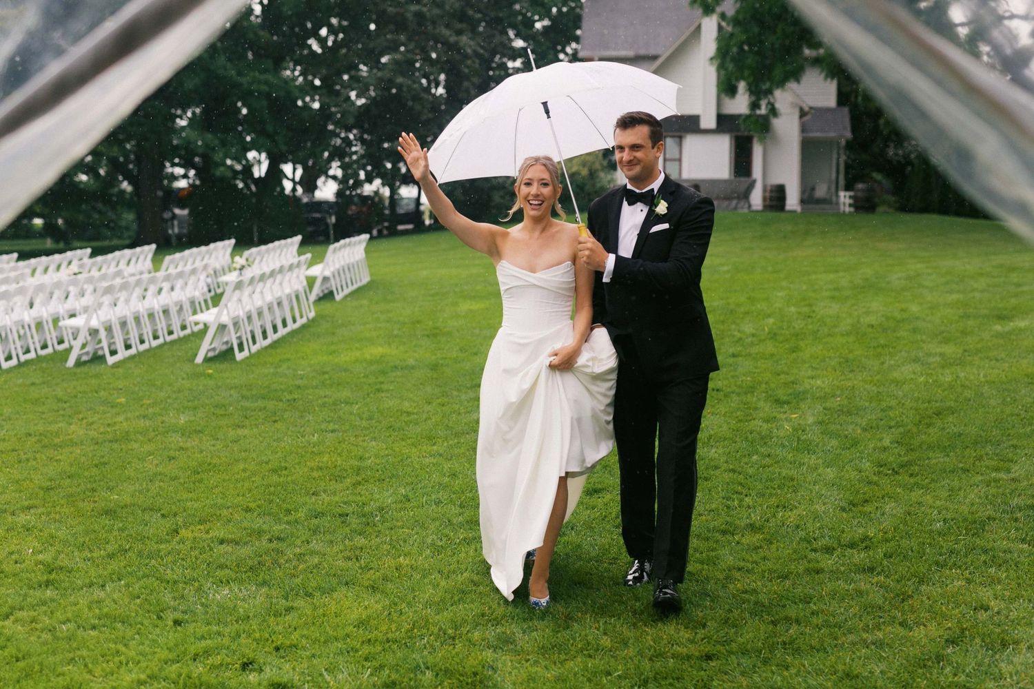 A couple celebrates their wedding day walking together through rain on a lush green lawn with white chairs set up for the ceremony.