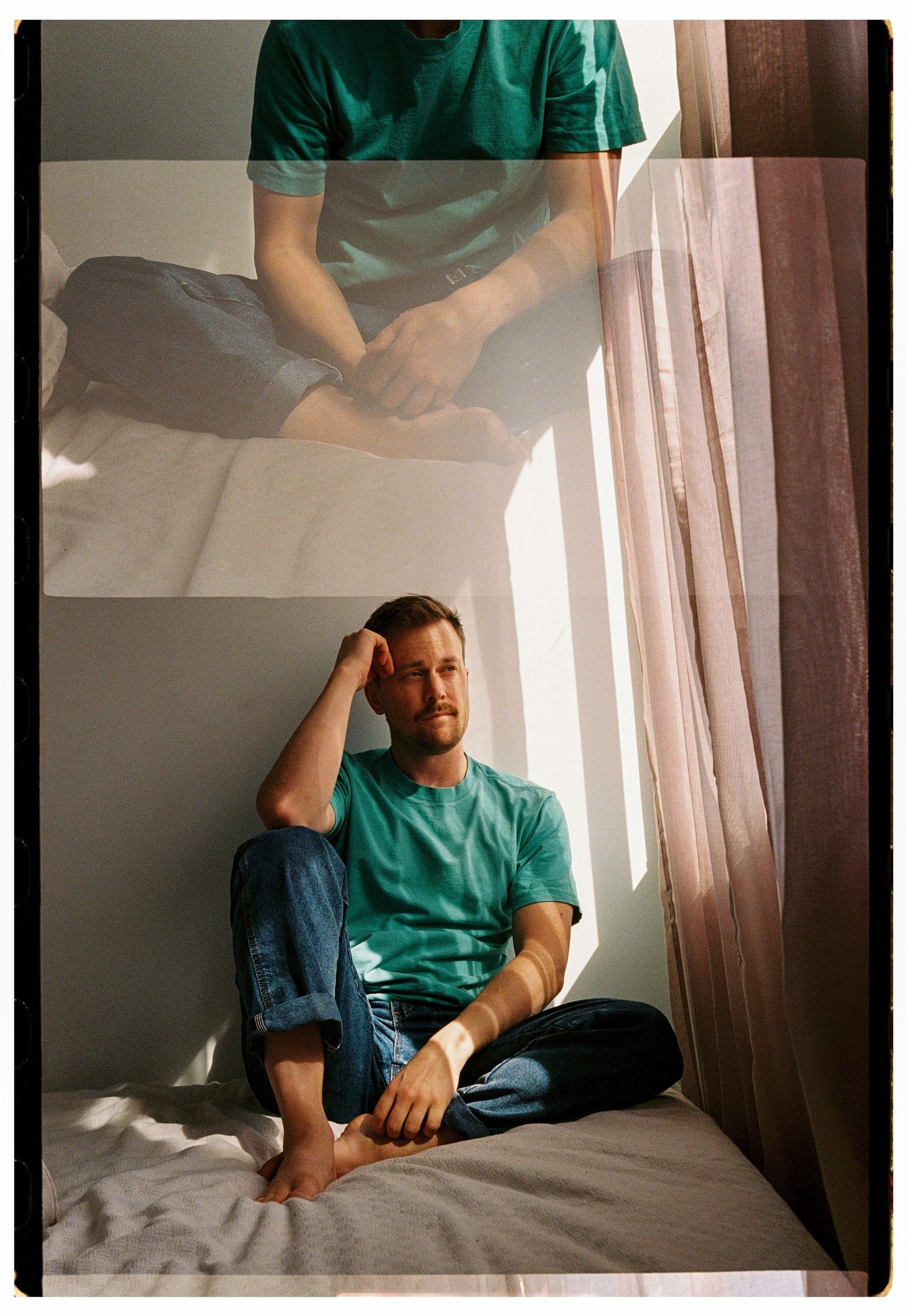 Man in teal shirt and jeans sits cross-legged on bed by sunlit window in double-exposure photo.