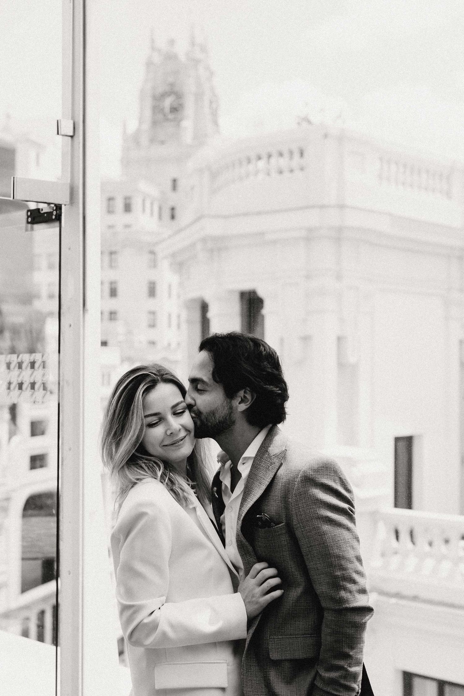 Black and white photo of a couple embracing on a balcony with a historic building backdrop.