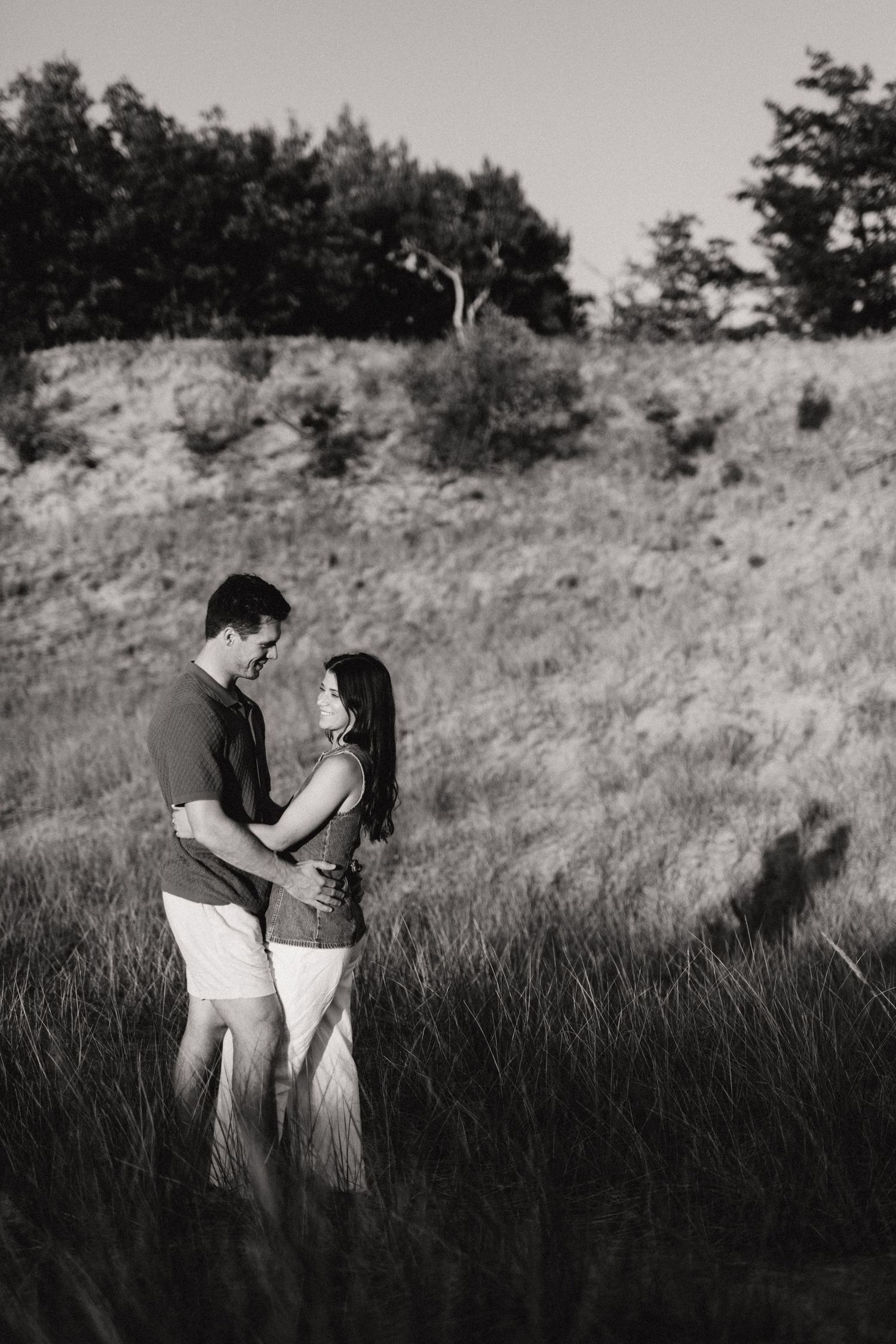 Black and white photo of a romantic couple embracing in a golden grass field at sunset.