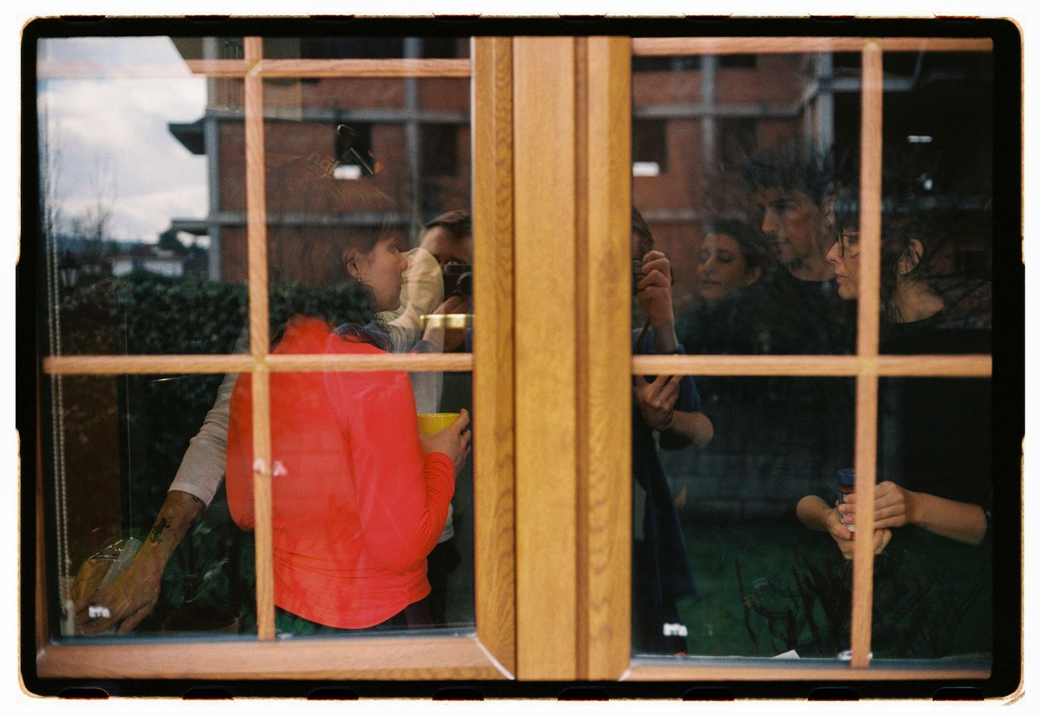 People gathered behind a wooden-framed window, one wearing a bright orange vest, viewed from outside.