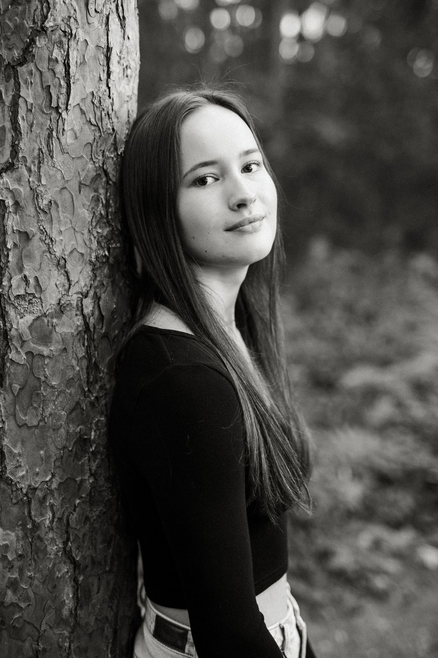 Black and white portrait of a teenage girl with long hair leaning against a tree trunk in a forest.