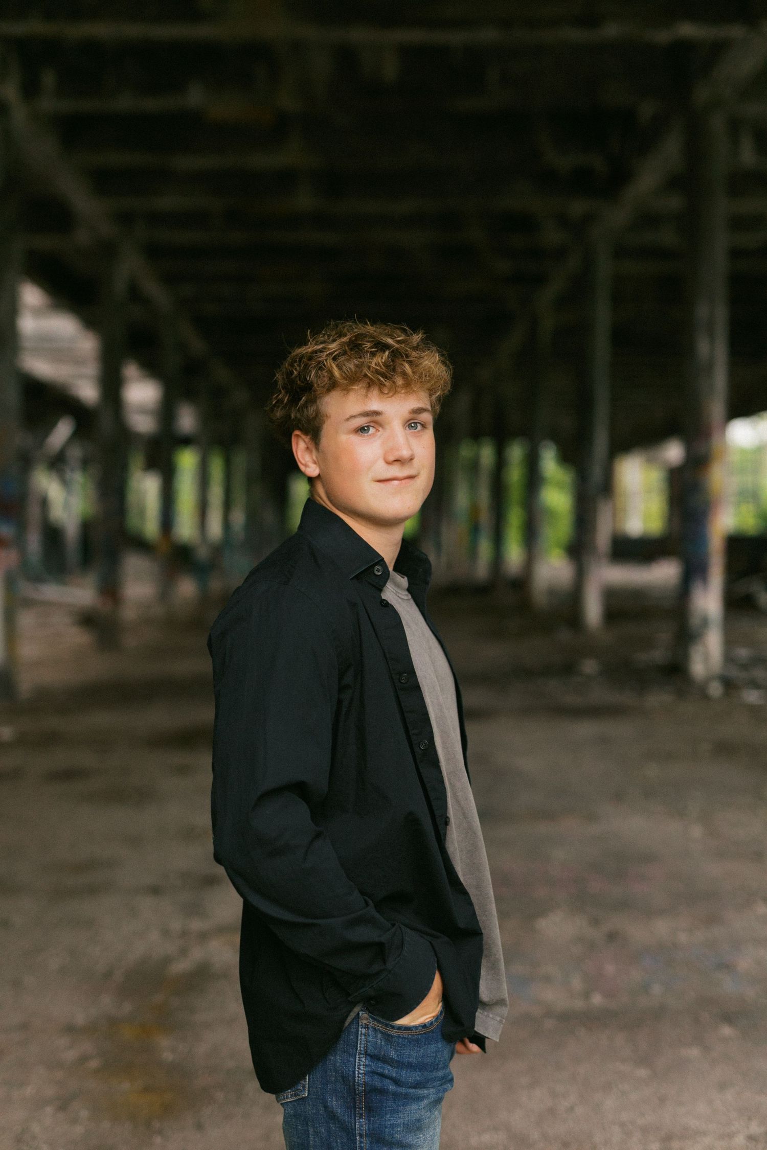 Teenage boy in black jacket and jeans posing in rustic industrial warehouse space.