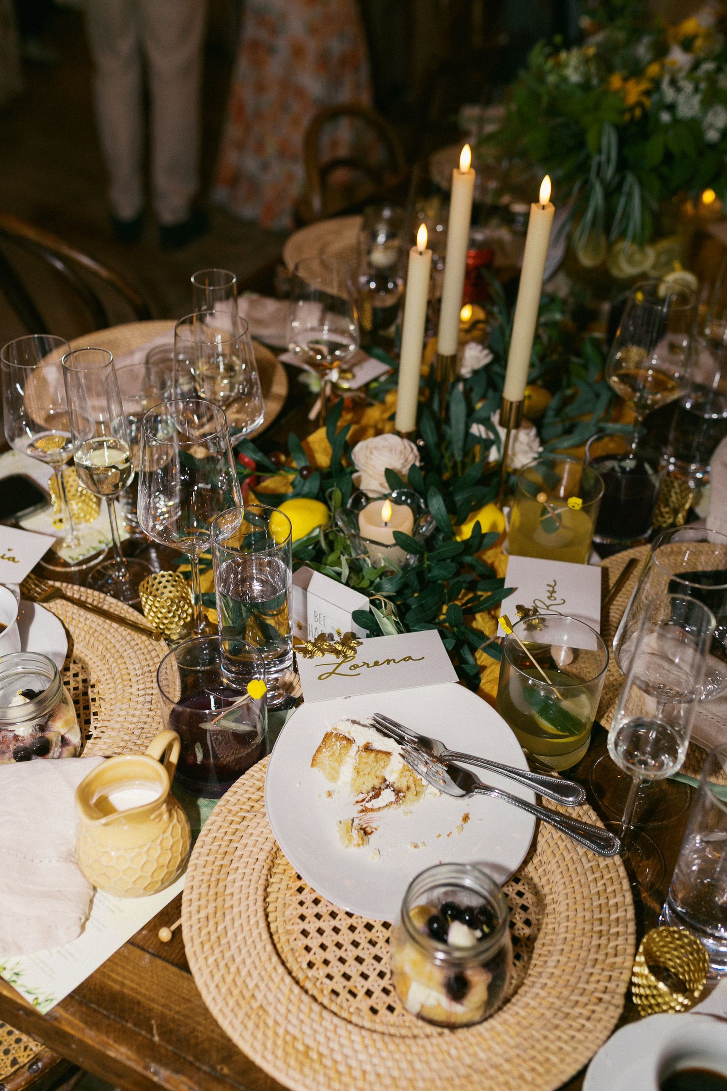 Elegant wedding reception table with candles, greenery, glassware, wicker placemats, and partially eaten cake slice.