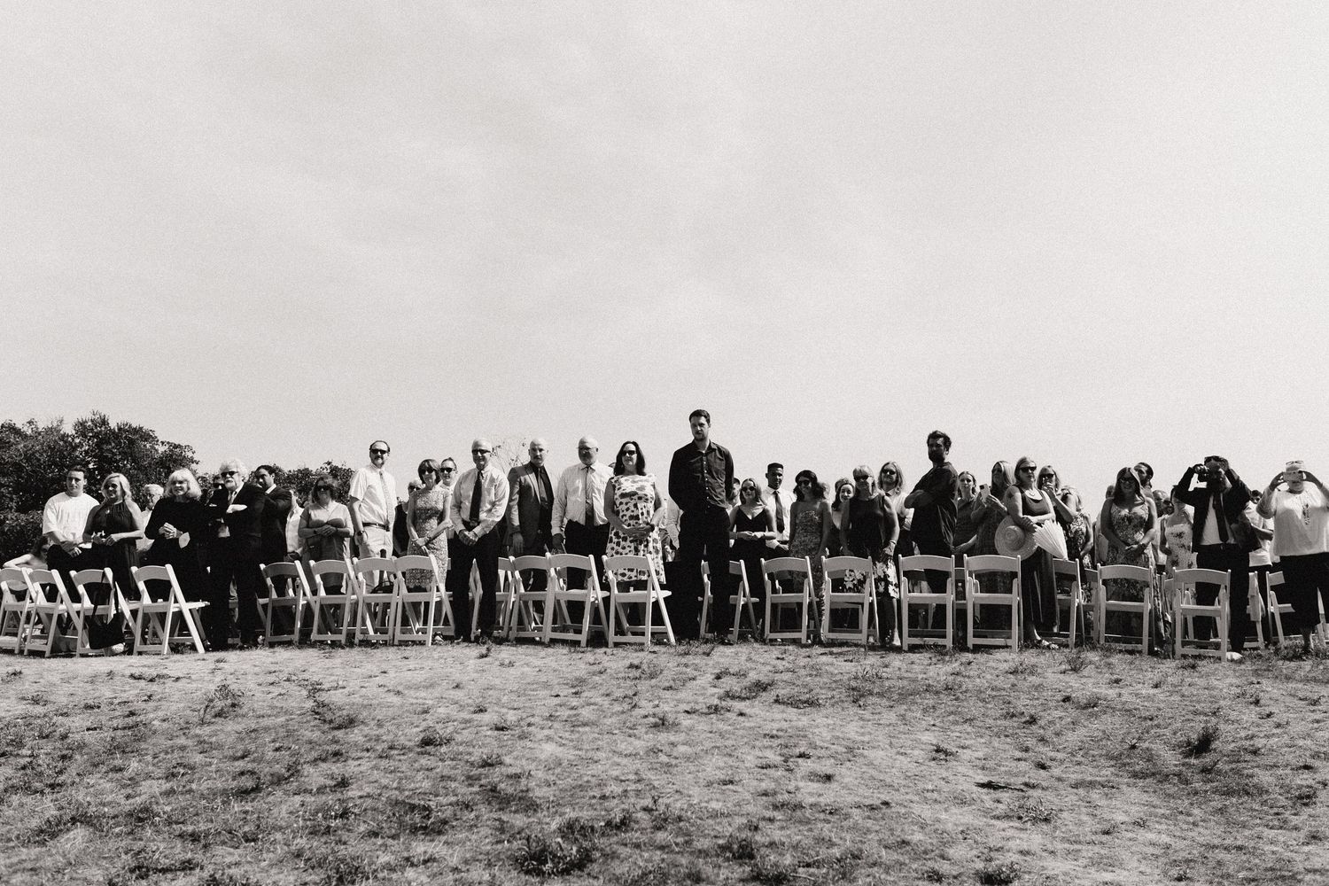 A black and white photo shows wedding guests seated in rows of chairs outdoors.