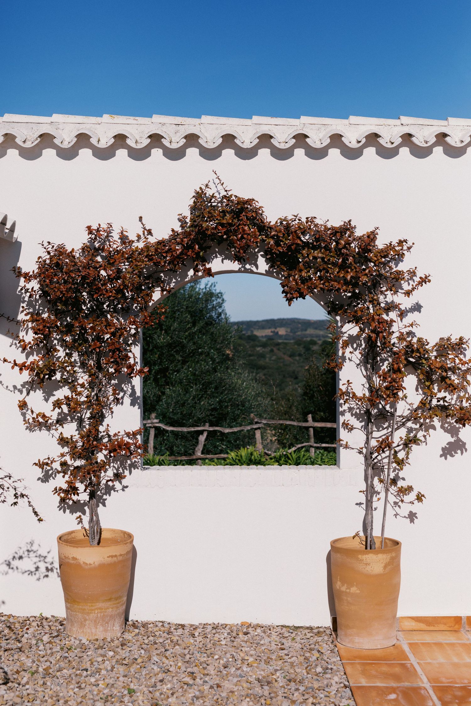 White Mediterranean wall with arched trellis, potted trees, and scenic countryside view through square window opening.