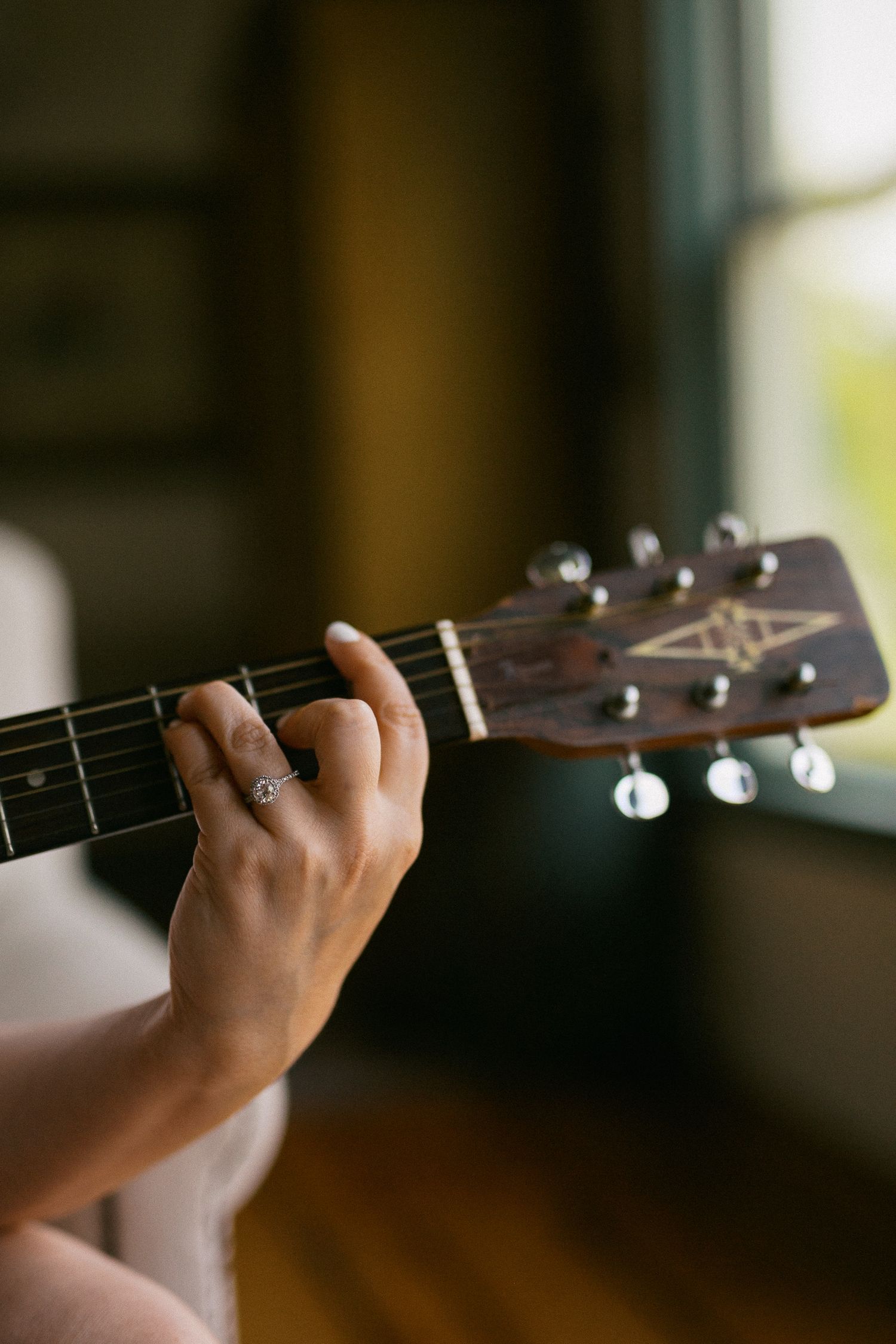Close-up of a hand with a ring playing chords on an acoustic guitar near a window.