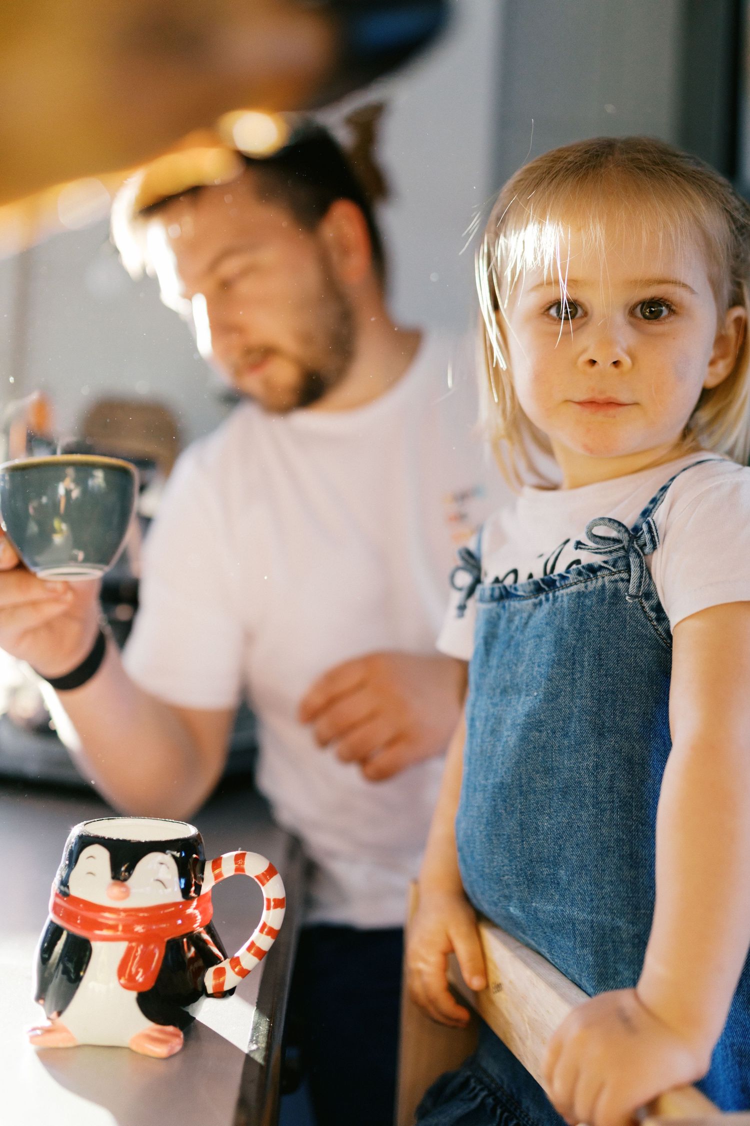 Young blonde girl sitting on kitchen counter beside bearded father holding a coffee cup.