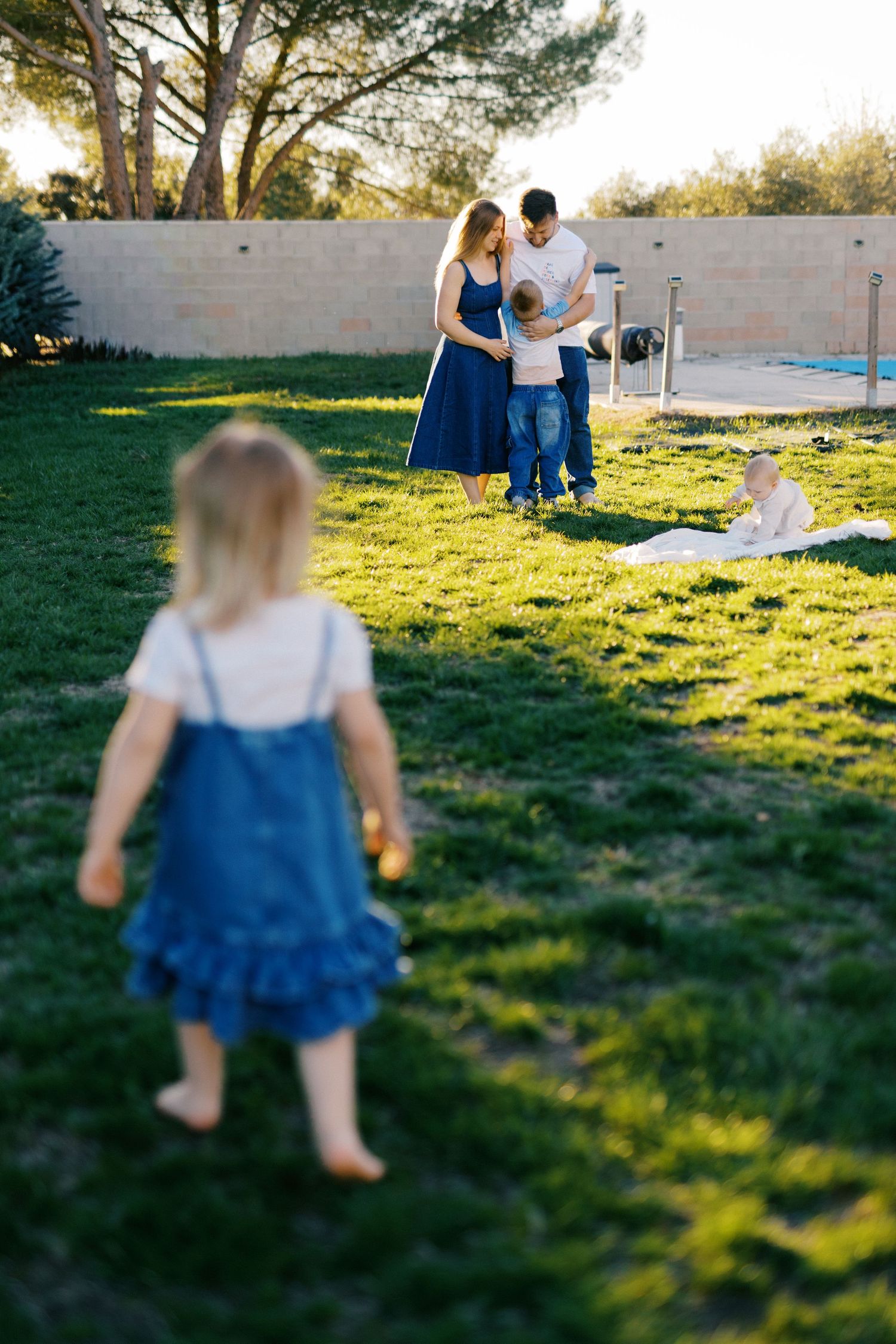 Family enjoying sunny backyard while toddler girl in blue dress walks toward parents.