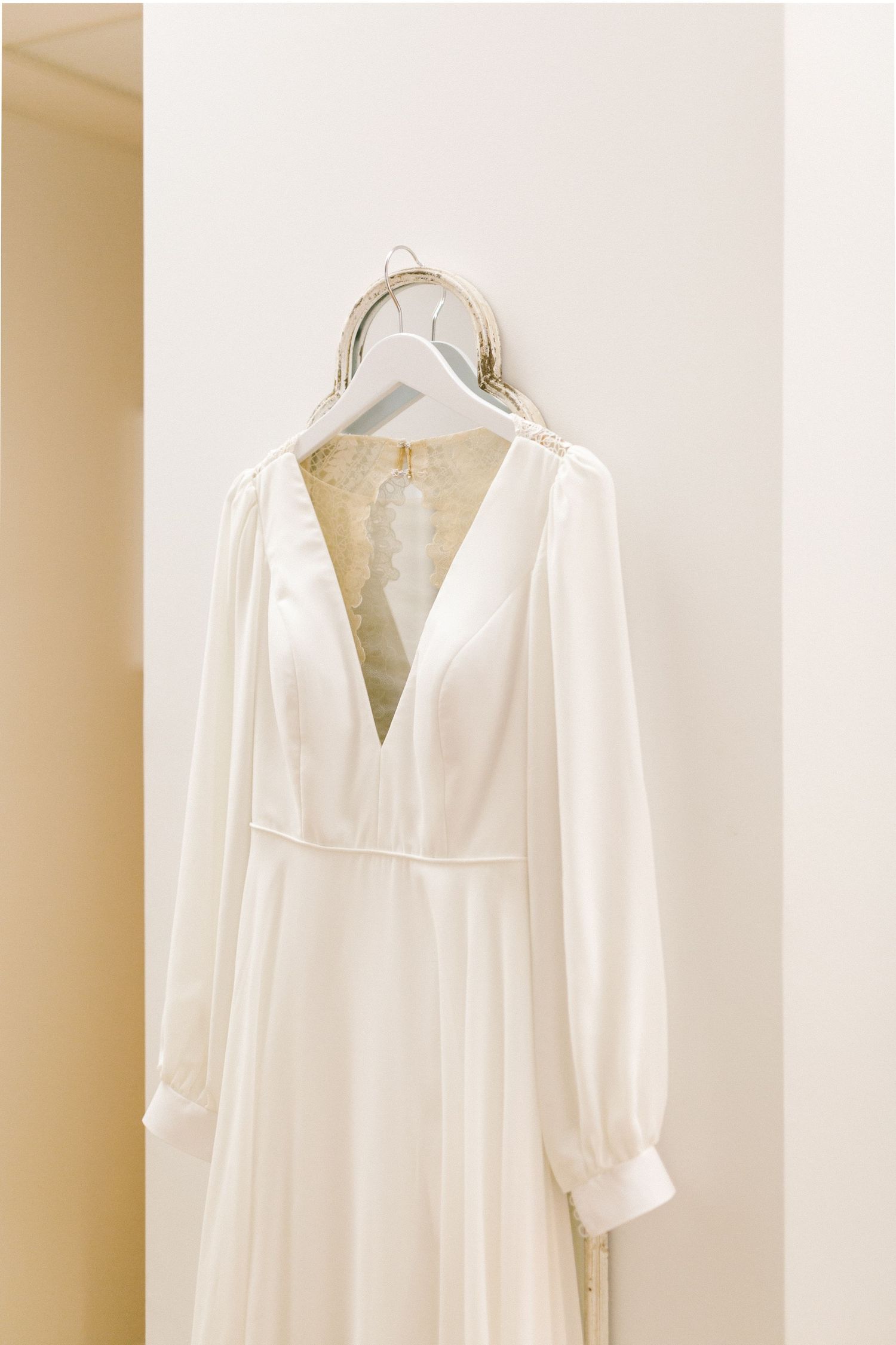 A classic white wedding dress with long sleeves and plunging v-neck hanging on a hanger against a beige wall.