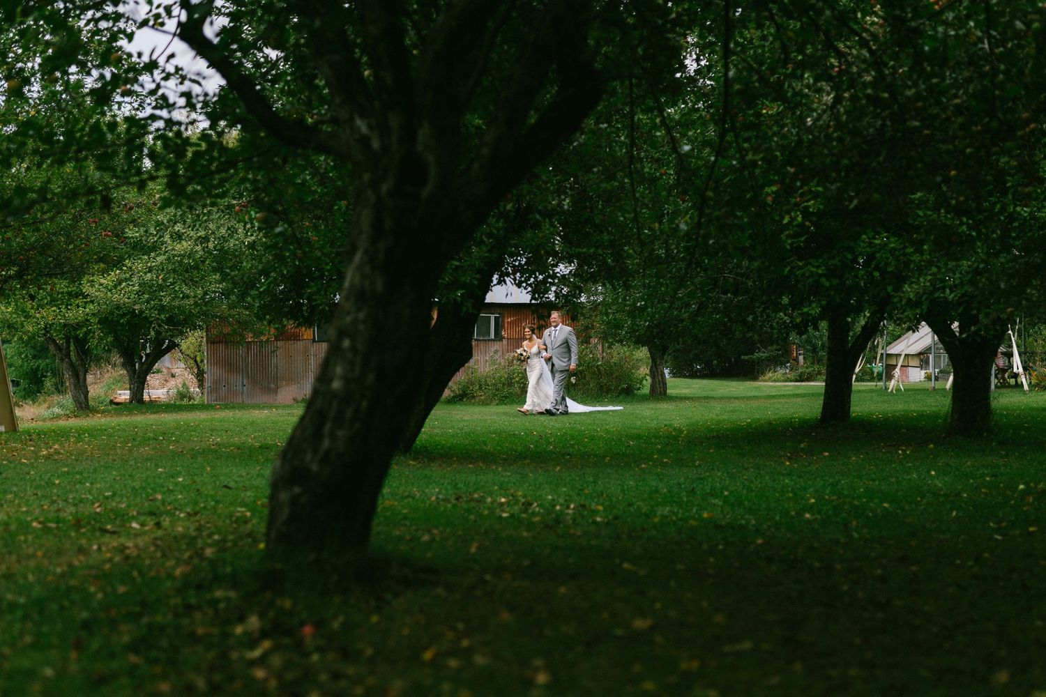 Bride and her father in a lush green park surrounded by mature trees with a white building visible in the background.