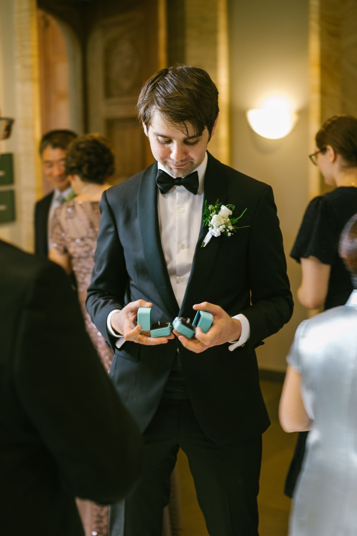 Groom in black tuxedo with bow tie and boutonniere holding two teal ring boxes at a wedding ceremony.