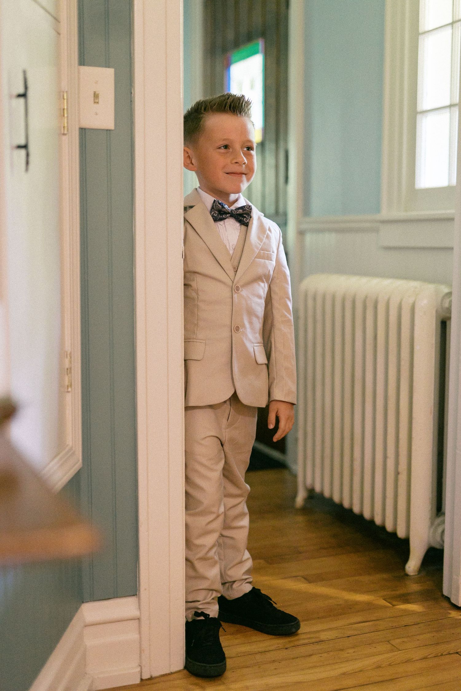 Young ring bearer in beige suit stands in hallway with white walls and wooden floors.