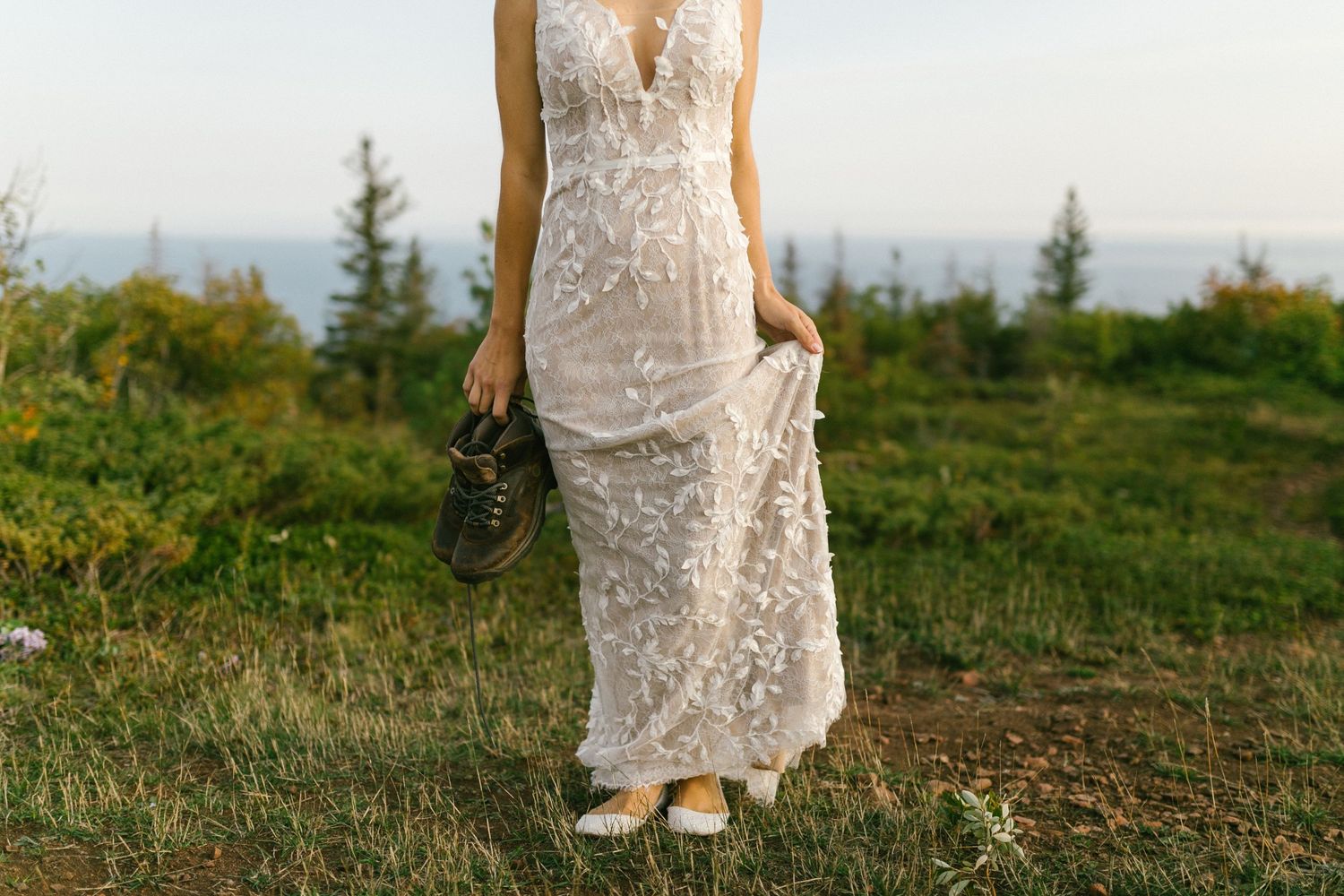 Close-up of a bride holding her lace wedding dress while walking on a grassy hilltop at sunset.