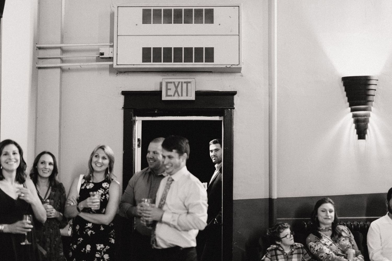 A group gathered by an exit door in a hallway with a wall sconce light fixture.