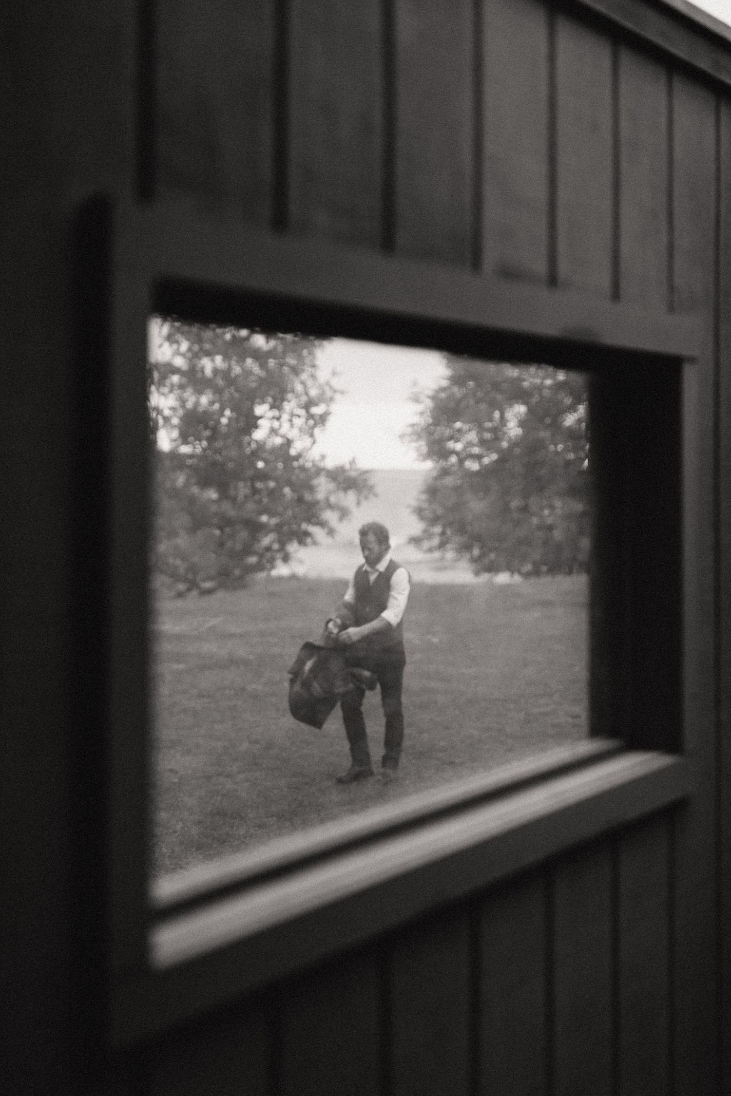 Black and white view through a window frame of a figure holding a bag in a grassy field.