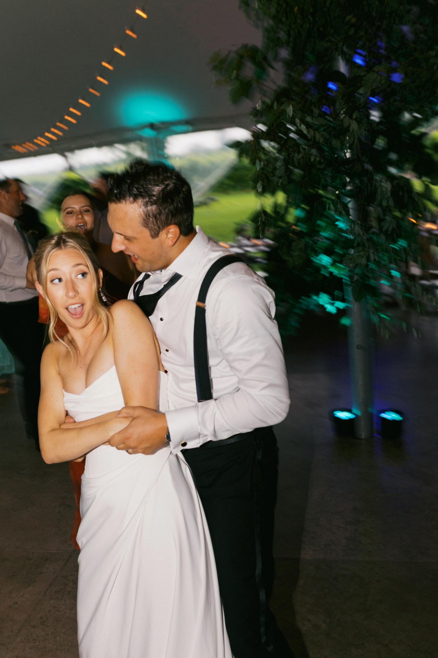 A couple in formal attire share a playful dance moment at an evening reception with colorful lighting.