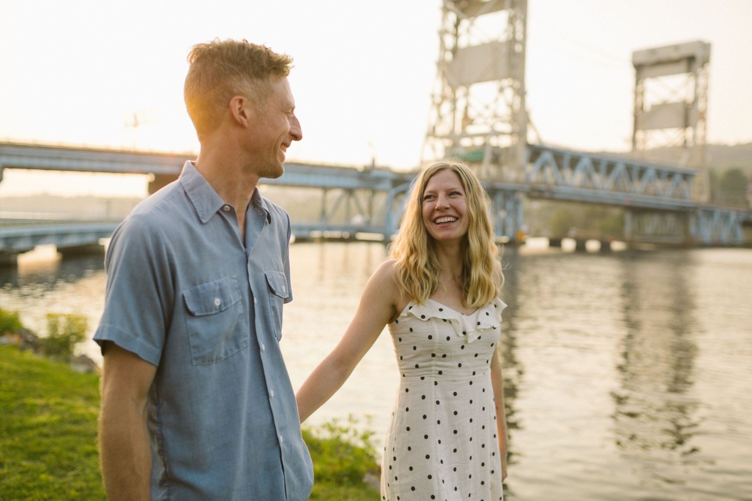 Couple laughing together near a bridge with one wearing polka dot dress and other in blue shirt.